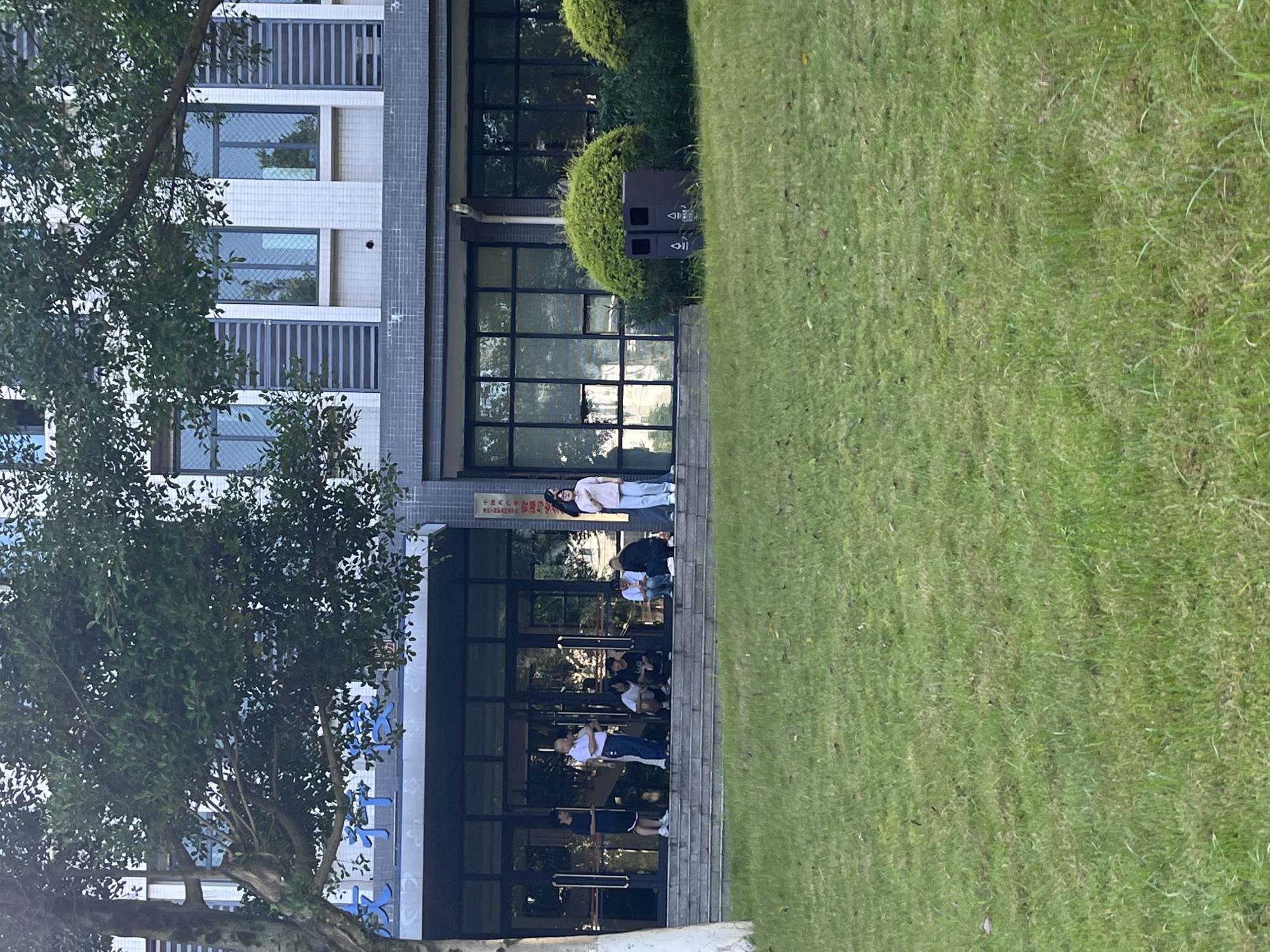 The image shows the exterior of a modern building, likely an educational institution, with a grassy lawn in the foreground. Several individuals are visible on the steps and near the entrance of the building.
In the foreground, a well-maintained lawn dominates the lower half of the image. The grass is green with some areas of drier, lighter-colored patches. A few fallen leaves are scattered across the lawn.
The midground features a set of concrete steps leading up to the entrance of a building. On these steps and just in front of the entrance, a group of approximately seven people are gathered.
One young person in a dark t-shirt and dark shorts stands on the far left, facing slightly left.
To their right, an older person in a white polo shirt and blue jeans stands talking to another individual who is mostly obscured.
Further to the right, several younger individuals are sitting on the steps in a relaxed manner. One is wearing a black t-shirt and shorts, another in a black t-shirt and dark pants, and a third in a dark top and jeans.
To the right of these seated individuals, a young person in a white t-shirt and light-colored pants stands, facing slightly to the left, with their hands at their sides.
Further to the right, near a trash can, a young person in a pink long-sleeved shirt and light-colored pants stands facing to the left.
The building itself has a modern architectural style with a facade of white tiles and dark gray horizontal banding. Large glass windows and doors form the entrance area. Above the entrance, blue Chinese characters are visible on the building, possibly indicating the name of the institution. The upper floors of the building feature rows of windows with dark frames.
To the right of the building entrance, two dark-colored trash receptacles are visible, each with a triangular recycling symbol. Behind the trash receptacles, lush green bushes are situated.
The overall environment suggests a daytime scene, likely in the late afternoon given the soft lighting. The presence of students and the setting imply a campus environment in Jiangjin District, Chongqing, China.