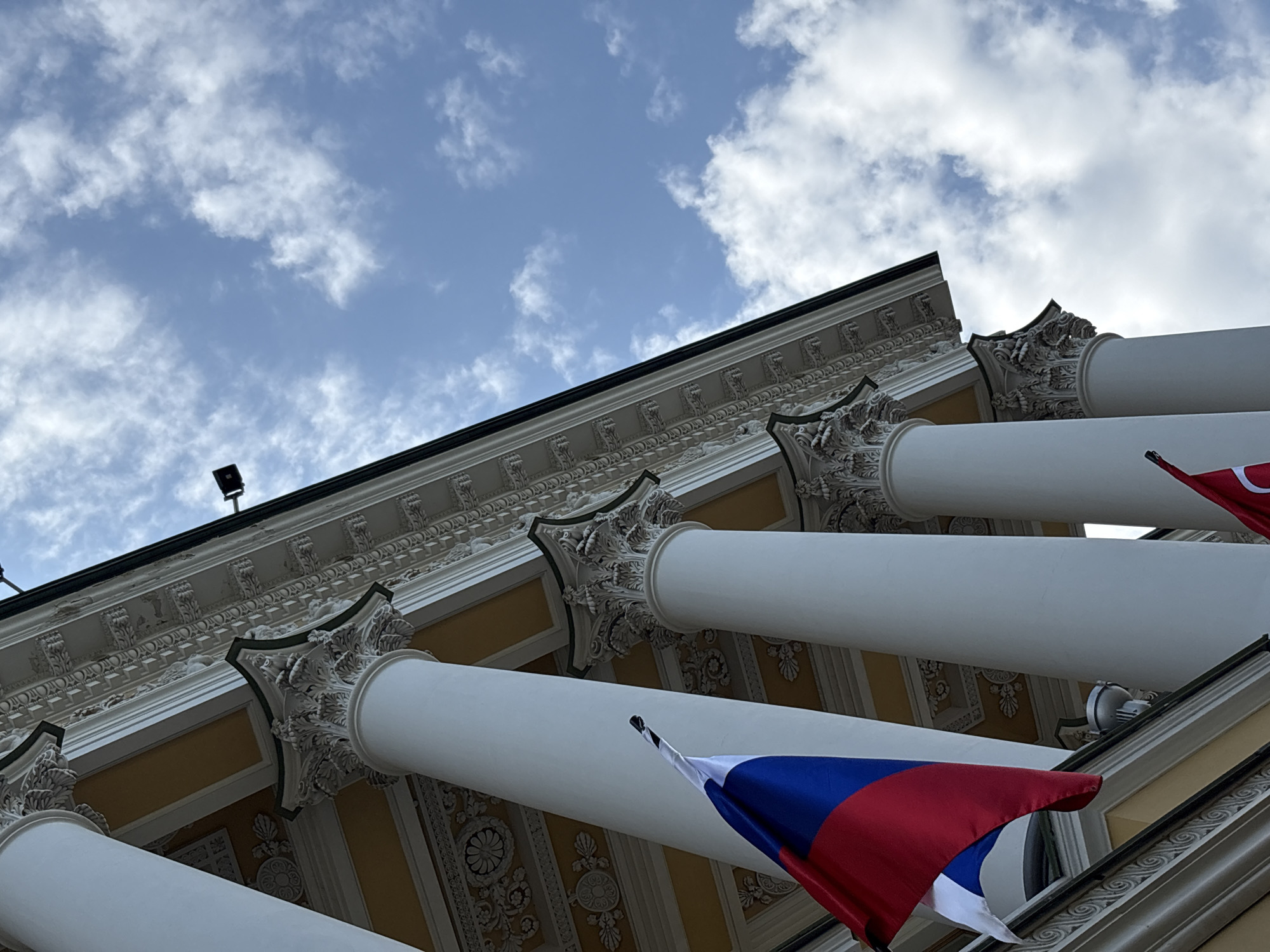 A low-angle shot captures the facade of a building in Saint Petersburg, Russia, under a partly cloudy blue sky. The building features classical architecture with white columns, ornate friezes, and a yellow-painted ceiling visible between the columns. A Russian flag, with its red, white, and blue horizontal bands, is prominently displayed in the foreground, attached to the building. Another flag, predominantly red with a white cross, is partially visible at the bottom right of the frame. Two dark security cameras are mounted on the building's exterior. The overall scene is static, with no people visible.