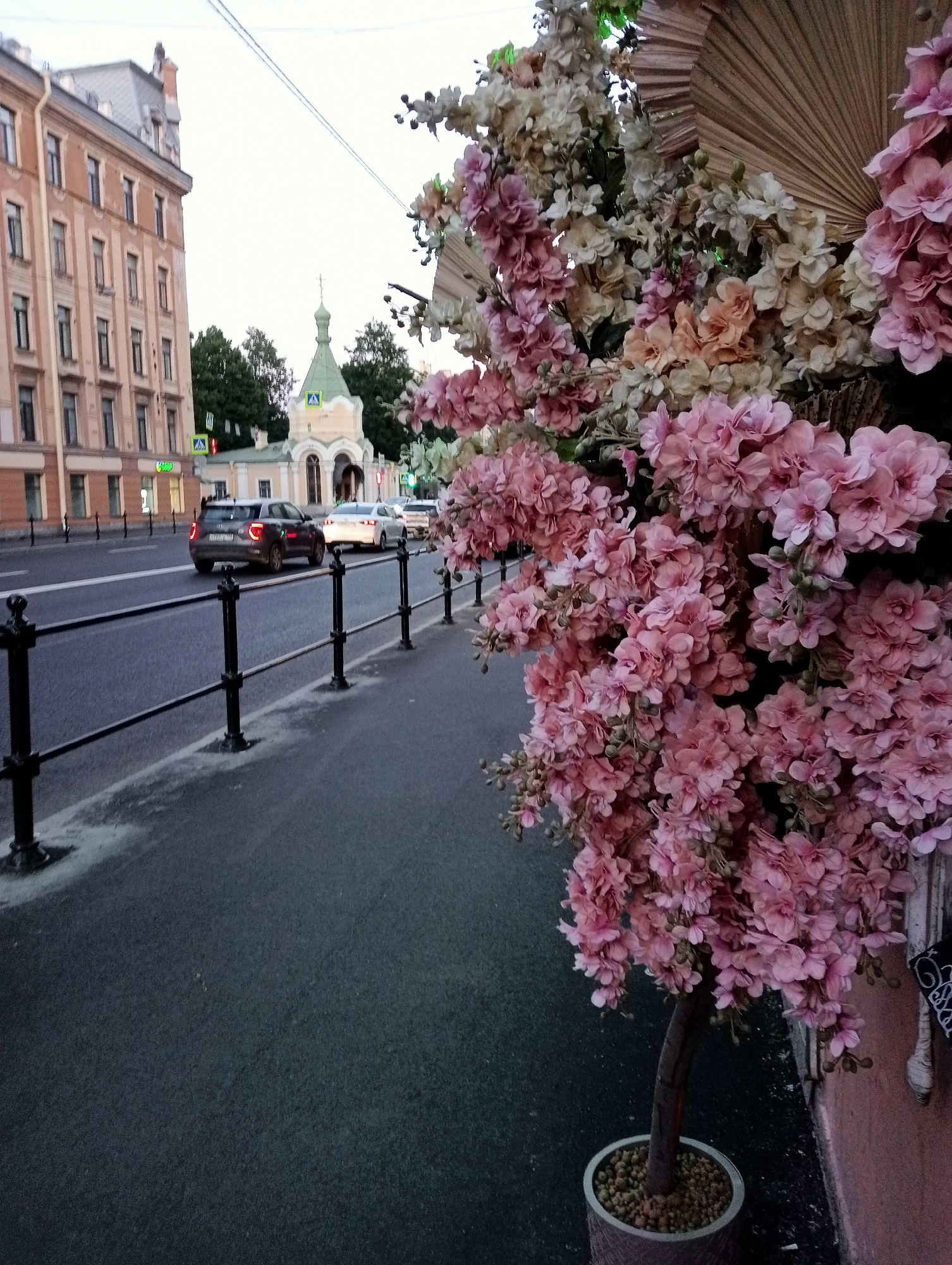 The image depicts a street scene in Saint Petersburg, Russia, near the Church of the Transfiguration of the Savior. A sidewalk is lined with decorative potted trees adorned with pink and white artificial flowers. A dark asphalt road runs alongside the sidewalk, with a black metal railing separating the two. Several cars are visible on the road, with one silver car and a dark grey car closest to the viewer. In the background, the pale yellow facade of a building with multiple windows and a green-domed church tower are visible. The sky is overcast, and the overall lighting suggests it is late afternoon or early evening.