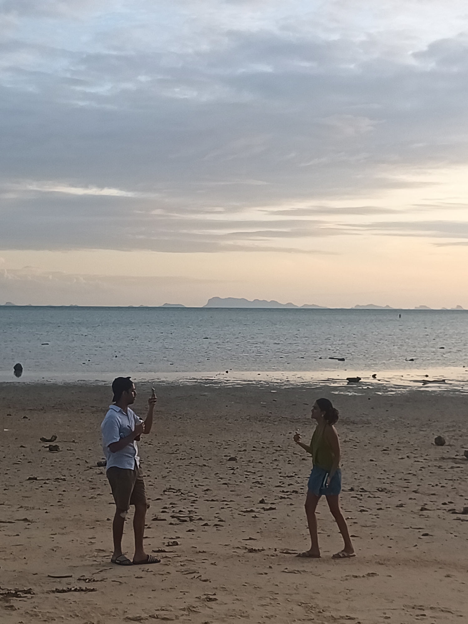 Two people are standing on a beach in ตำบลเกาะพะงัน, Thailand. The person on the left is a man wearing a light blue short-sleeved shirt, brown shorts, and sandals. He is holding a smartphone in his right hand, appearing to take a photo. The person on the right is a woman wearing a green tank top and denim shorts. She is looking to her left and holding a small object in her right hand, possibly also a phone. The beach is sandy with some scattered debris. The ocean is calm and stretches to the horizon, where several islands are visible in the distance. The sky is overcast with soft, muted colors, suggesting it is either early morning or late evening.