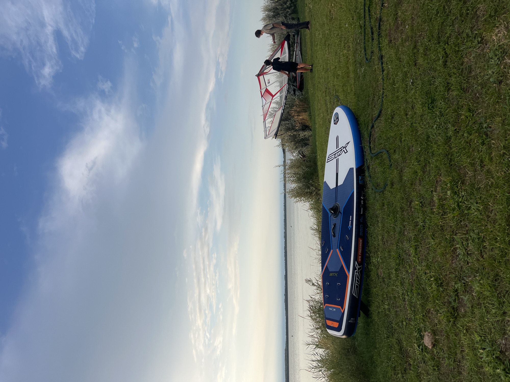 In the city of Am Salzhaff, Germany, two individuals are by the water, preparing for windsurfing. In the foreground, a blue and white paddleboard lies on the grass. Further back, a red and white windsurfing sail is propped up, with two people standing near it. The scene is set by the water, with a calm lake or sea in the background, and a cloudy sky overhead. The overall atmosphere suggests an outdoor recreational activity.