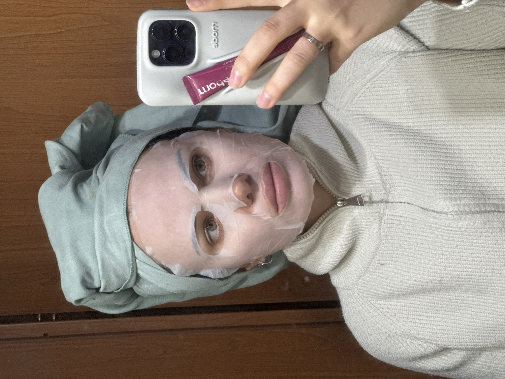 A female individual is depicted in a mirror selfie taken indoors in Belgrade, Serbia. She is observed from the waist up, facing the camera indirectly through a reflection. Her face is covered with a white sheet face mask, exposing her eyes, nose, and mouth. Her head is wrapped in a light green towel. She wears a cream-colored, full-zip, knitted sweater. Her left hand, adorned with a silver-colored band ring on the ring finger, holds a light grey smartphone with a visible triple camera array. A small magenta-colored tube, displaying reversed text including "LUXIOUS" and "ehori", is held against the phone case, which also shows reversed branding "eborh". The background consists of a light brown wooden surface, possibly a door or wall panel, with visible vertical lines and wood grain. The subject's visible right eye is light brown. The lighting is consistent with an indoor environment.