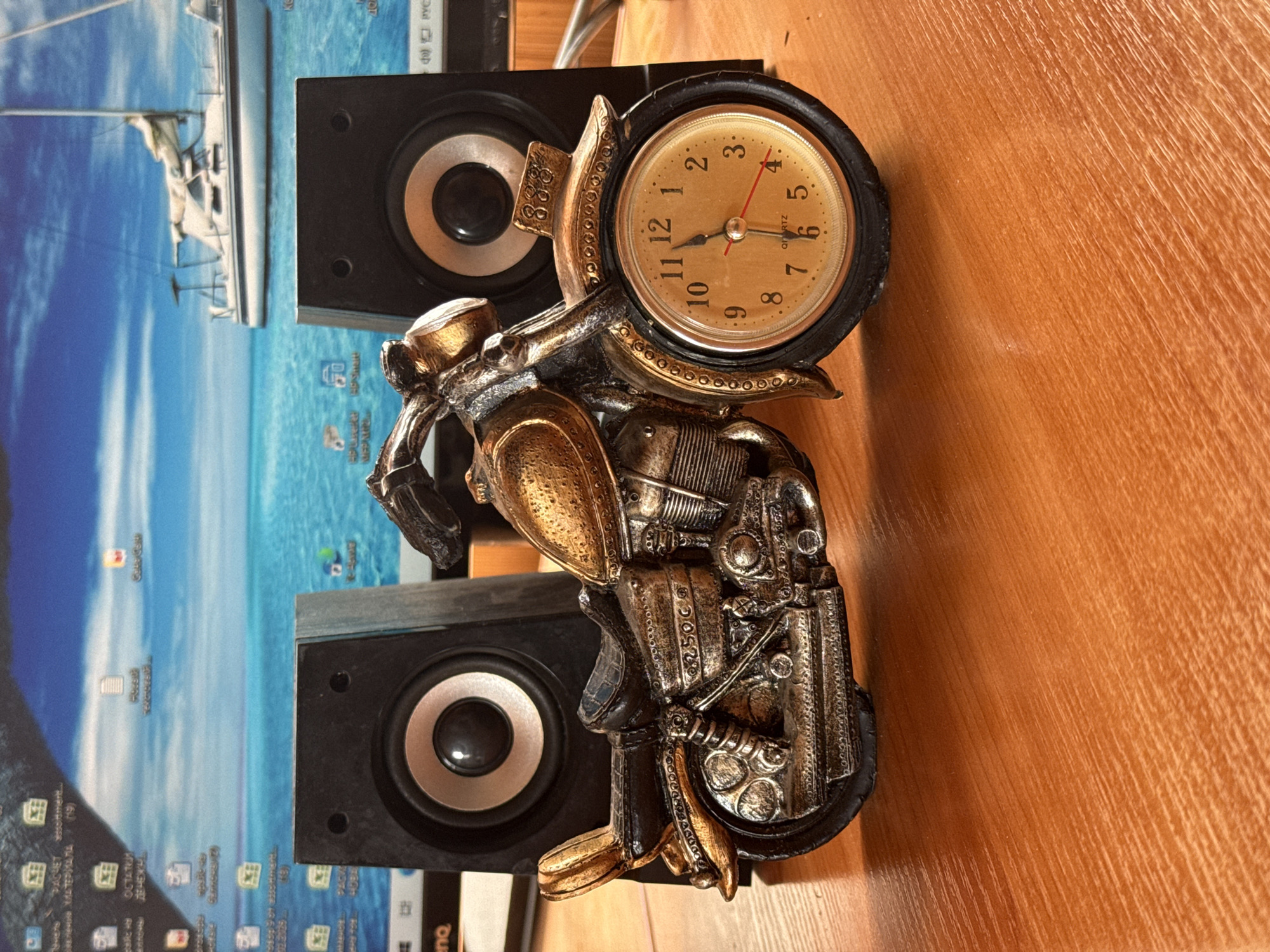 A decorative motorcycle clock is displayed on a wooden surface in front of two black speakers. The motorcycle is golden and appears to be made of metal, with intricate details on the engine, wheels, and seat. The clock is integrated into the front wheel of the motorcycle and has a golden face with black numbers and hands. The background shows a computer monitor displaying a desktop wallpaper of a sailboat on a blue ocean, with various icons visible on the screen. The overall scene suggests a personal workspace or desk setup.