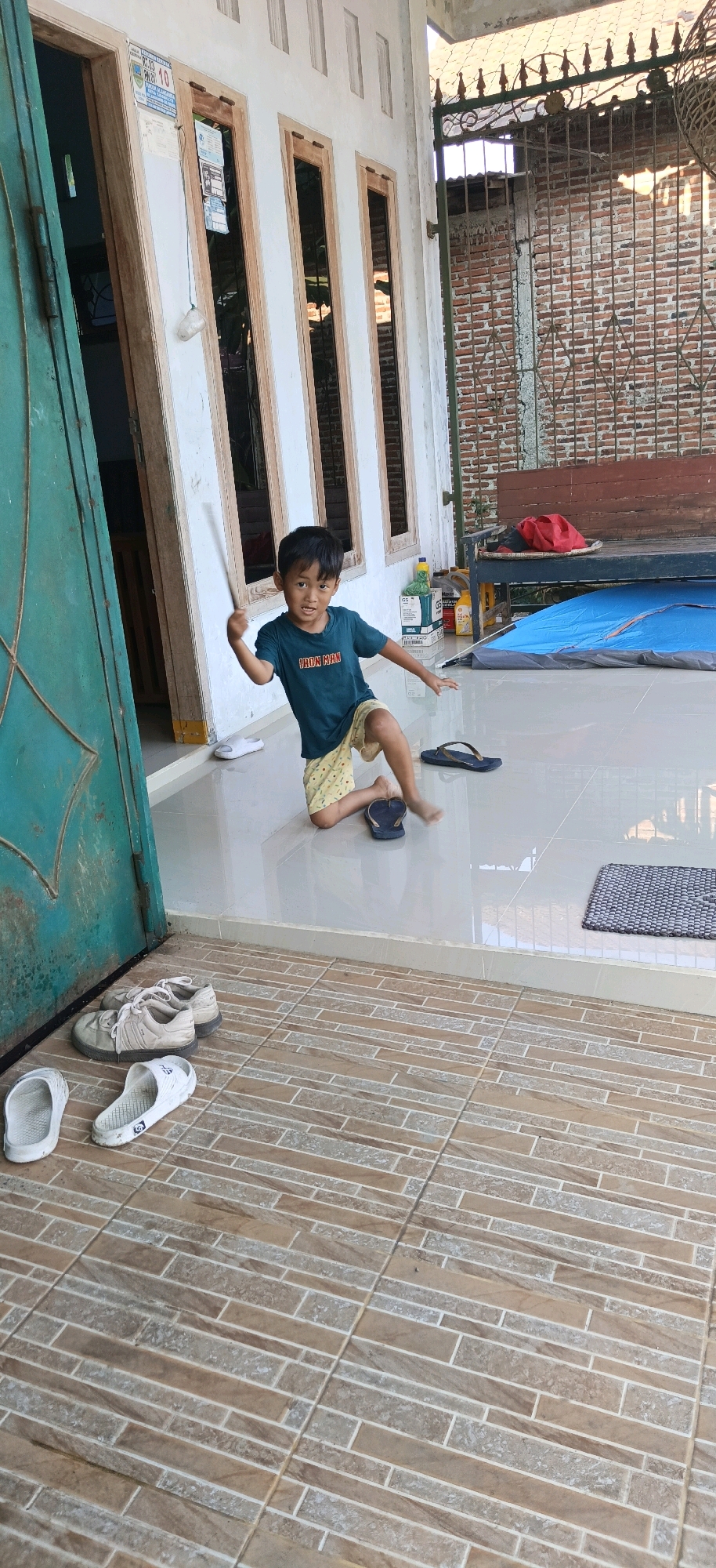 A young boy kneels on a tiled porch, holding a thin stick. He wears a dark blue t-shirt with "IRON MAN" printed on it and yellow shorts with a checkered pattern. The boy is positioned in front of a white-walled building with wooden-framed windows. To his left is a green door. Several pairs of footwear, including white flip-flops and tan sneakers, are scattered on the floor in the foreground. Behind the boy, a blue tent is visible, along with a wooden bench and a brick wall. The scene appears to be outdoors, possibly in the city of Panjatan, Indonesia, given the architecture and general environment.