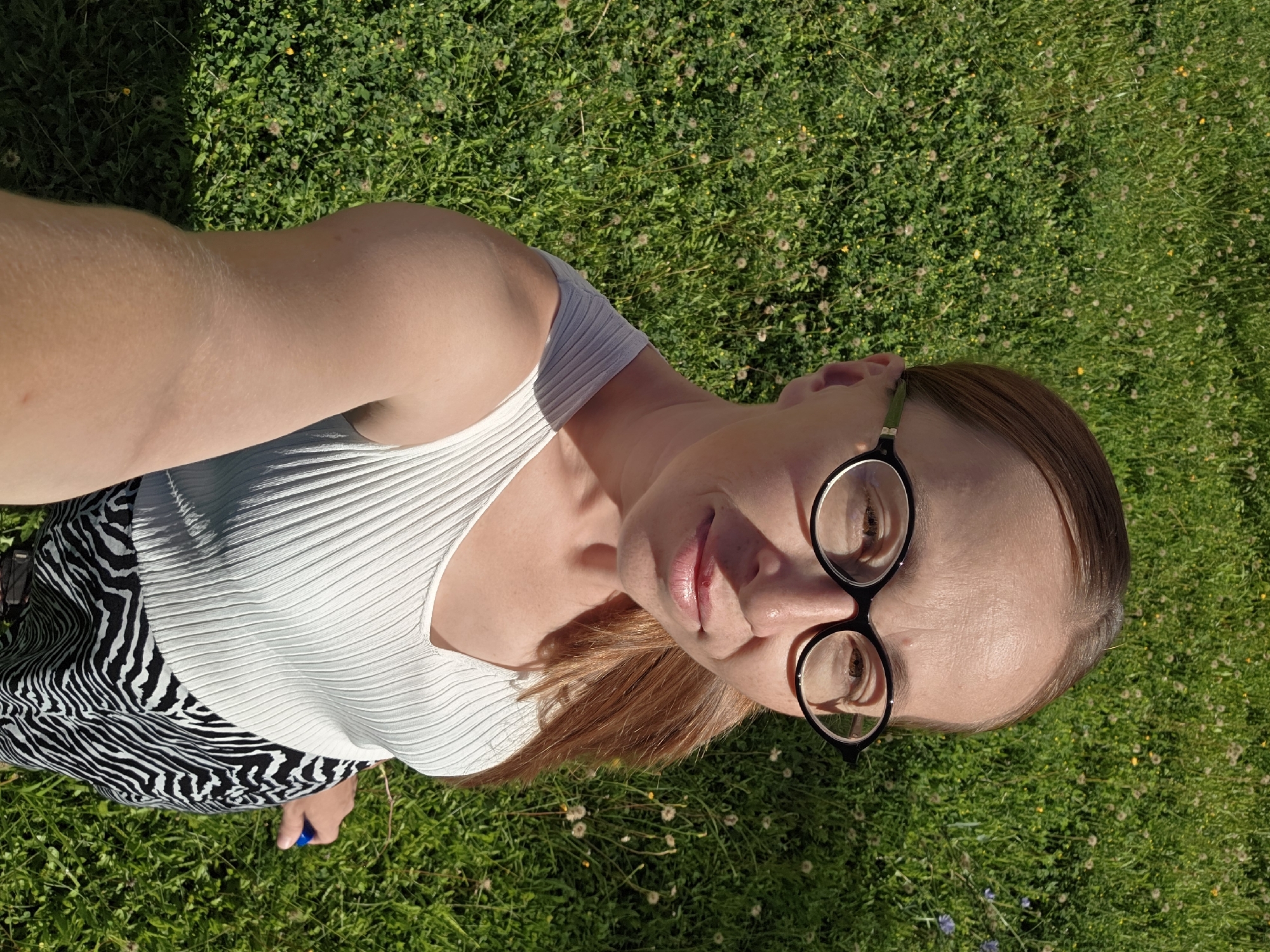 A high-angle, full-body selfie is captured outdoors on a sunny day, featuring a woman standing in a grassy area. The woman, with fair skin and reddish-brown hair pulled back into a ponytail, wears black-rimmed glasses, a white ribbed tank top, and a black and white zebra-print skirt. Her left arm is extended towards the camera, holding the phone for the selfie. The background consists of lush green grass dotted with small white and yellow flowers, typical of a meadow or park. The sunlight casts shadows on the grass, indicating a bright, clear day. The scene is consistent with an outdoor setting in Chaikovsky, Russia, likely during warmer months.