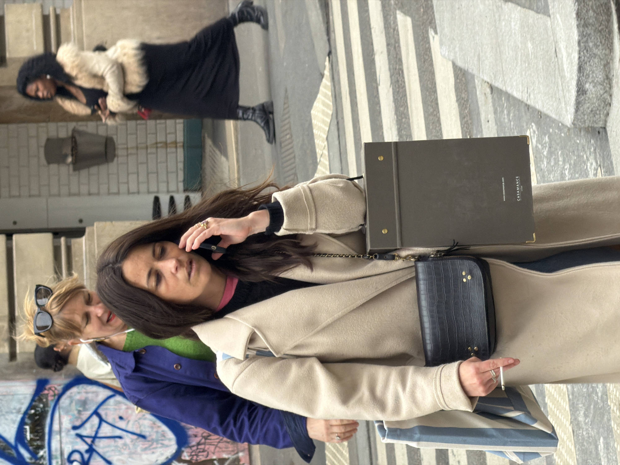 An urban street scene in Paris, France, features multiple pedestrians. In the foreground, a woman with long dark hair wears a beige trench coat over a black and pink top. She holds a black smartphone to her right ear, appearing to be on a call, while her left hand holds a lit cigarette, displaying a silver ring on her ring finger. She carries a black faux-croc shoulder bag, a blue and white striped tote bag, and a dark grey portfolio-style object branded "CASAMANCE." Behind her, a blonde-haired woman wears a navy blue jacket over a green top, with sunglasses pushed onto her head and white wired earphones in her ears, smiling towards the right. Partially visible behind her is another person with dark hair. In the background, a Black woman with curly dark hair walks from right to left, wearing a light beige fur-trimmed jacket over a long black dress and black boots, carrying a small red bag and holding a dark object. The environment includes a paved street with a white striped pedestrian crossing, a building with white tiled walls and a recessed grey fixture, and a graffiti-covered wall featuring prominent blue lettering, including "RAT." The scene is illuminated by daylight.
