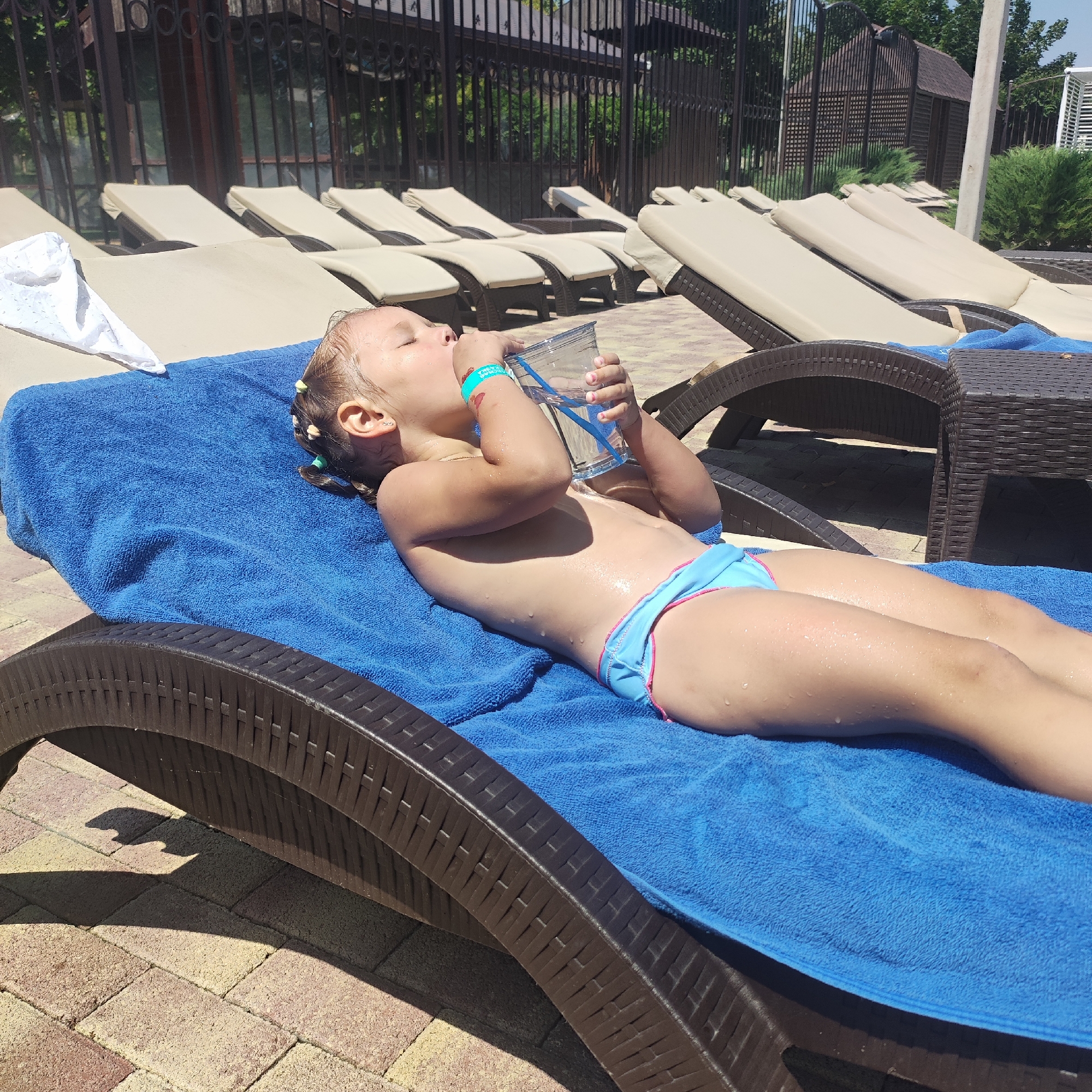 A young girl with blonde hair in pigtails lies on a blue towel on a brown lounge chair. She is wearing blue bikini bottoms. She is drinking from a clear plastic cup with a blue straw. Water droplets are visible on her skin. In the background, there are more lounge chairs, a fence, trees, and buildings. The sun is shining, indicating it is daytime. The scene appears to be at a pool or resort. The girl's eyes are closed as she drinks.