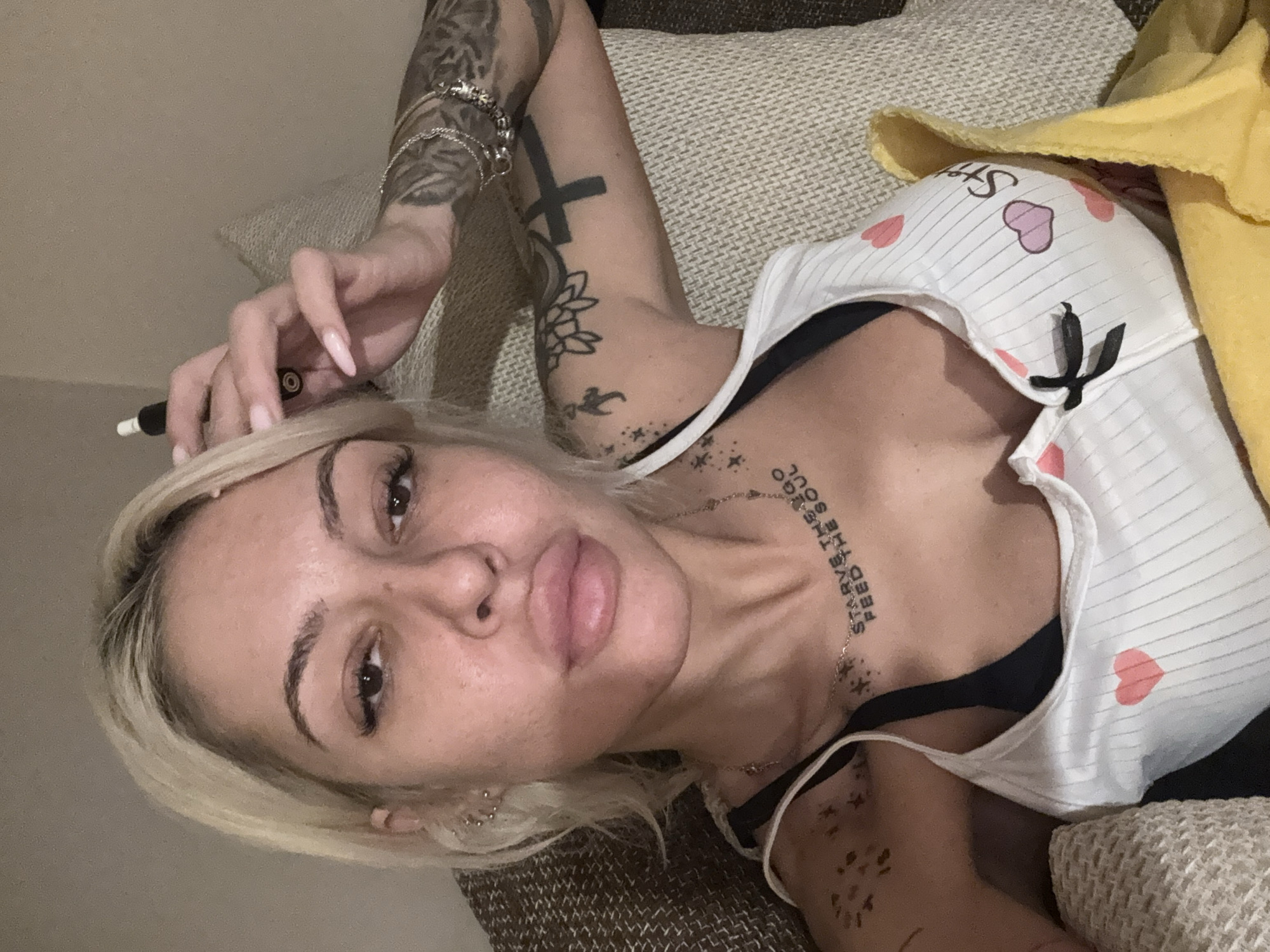 A woman with blonde hair and multiple tattoos is lying on a textured couch. She is wearing a white tank top with hearts and black trim, exposing a black bra. Her left arm is raised, holding what appears to be a vape pen. The background includes a yellow fabric draped over the couch. The image does not provide information about the city of Plovdiv, Bulgaria.