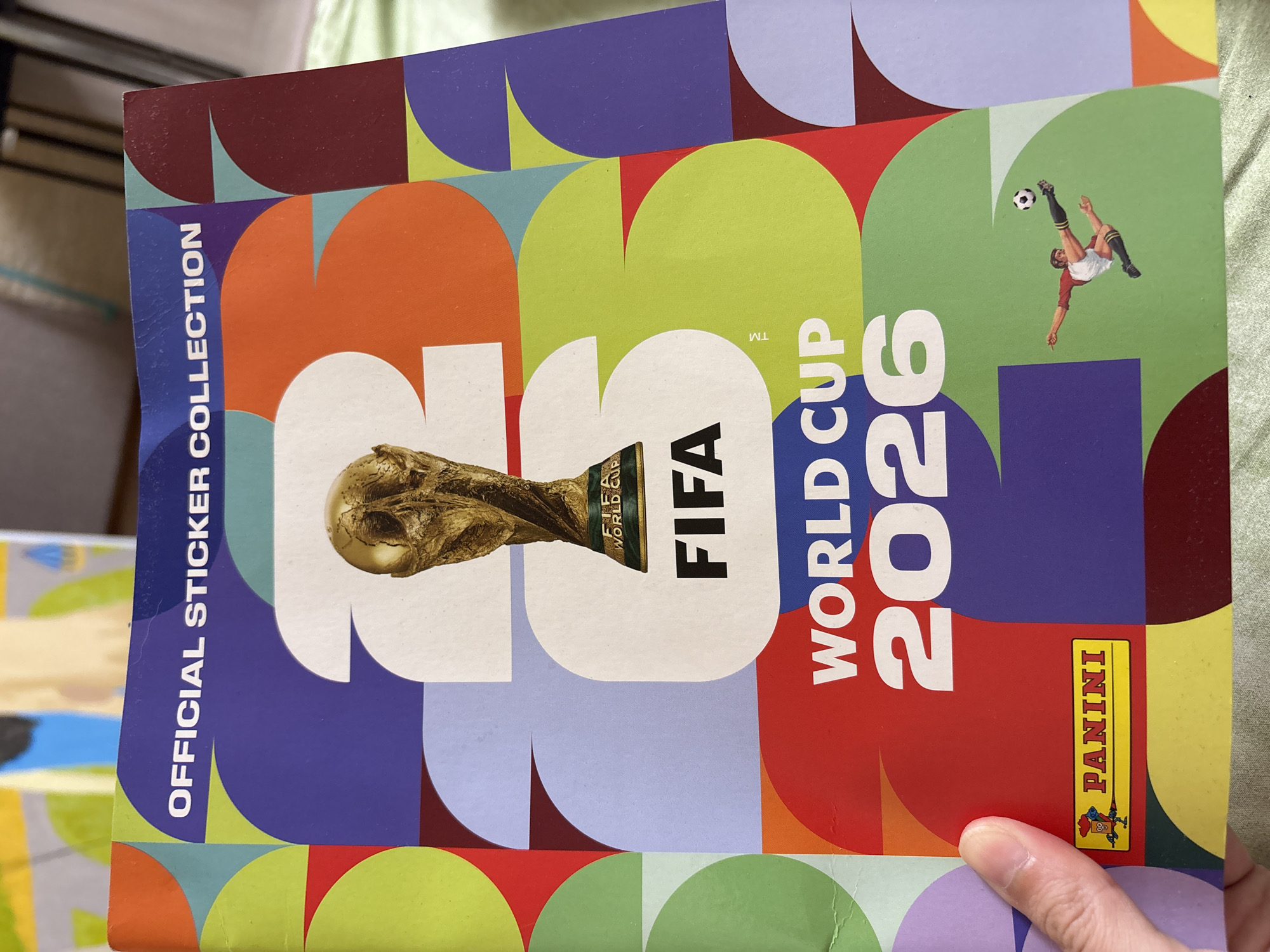 The image shows the cover of the Official Sticker Collection for the FIFA World Cup 2026. The cover features a graphic design with large, colorful shapes and the FIFA World Cup trophy prominently displayed. The words "OFFICIAL STICKER COLLECTION," "FIFA WORLD CUP," and "2026" are clearly visible. A small Panini logo is located in the bottom left corner. A stylized illustration of a soccer player kicking a ball is on the right side of the cover. The cover appears to be lying on a green, textured surface, possibly a bedspread. There is no information in the image that allows for determining a location in the city of Ветлужский, Russia.