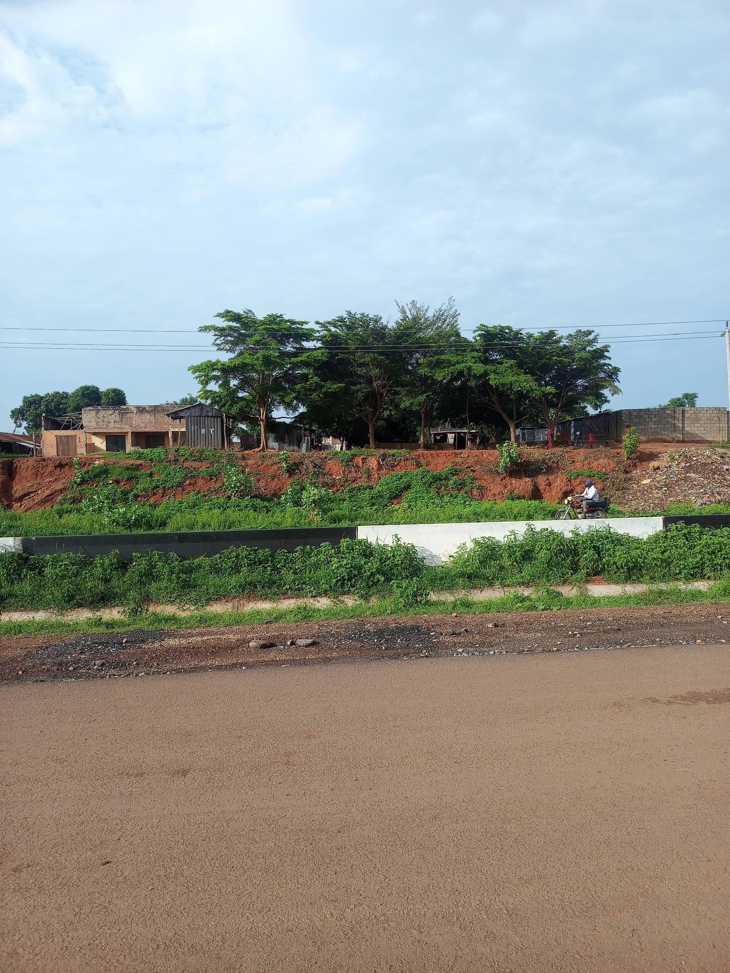 The image depicts a street scene in Keffi, Nigeria, with a focus on a section of elevated ground overlooking a road. The foreground is dominated by a paved road surface, appearing somewhat worn and textured. Immediately beyond the road is a strip of rough ground with scattered stones and reddish-brown soil, transitioning into a dense green growth of vegetation. This vegetation is contained by a concrete barrier, painted white and black.
The midground shows a steep embankment covered in lush green grass and some scattered smaller plants. The embankment is made of reddish-brown soil, indicating a significant cut into the landscape. On top of this embankment, a cluster of trees with full, green canopies provides shade. Beneath the trees, several structures are visible. To the left, there is an older building with what appears to be crumbling walls and a corrugated iron roof. Next to it is a small, dark, metallic shed. Further to the right, there are more structures with corrugated roofs and some laundry hanging to dry. A man is riding a motorcycle in front of these structures, wearing a white shirt and a blue cap. The ground behind the structures is a mix of red soil, rubble, and some discarded items, with a brick wall visible in the far right background. The sky above is partly cloudy, with a light blue hue. Overhead power lines are strung across the upper portion of the image.