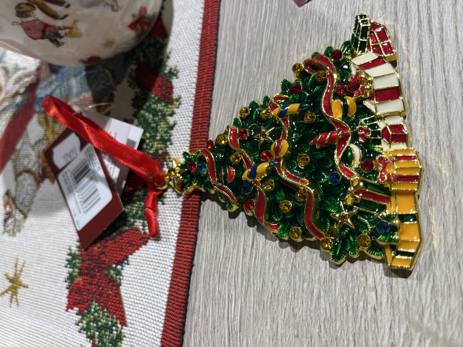 A Christmas tree-shaped ornament sits on a gray wood surface with a holiday-themed tablecloth to the side. The ornament is intricately decorated with gold trim, green enamel, and colorful rhinestones, resembling a Christmas tree adorned with lights, ribbons, and stars. At the top are stacked gift boxes. A red ribbon and tag are attached to the tree’s base. The tablecloth features a white background with a pattern of red poinsettias and green wreaths, accented by a red border.