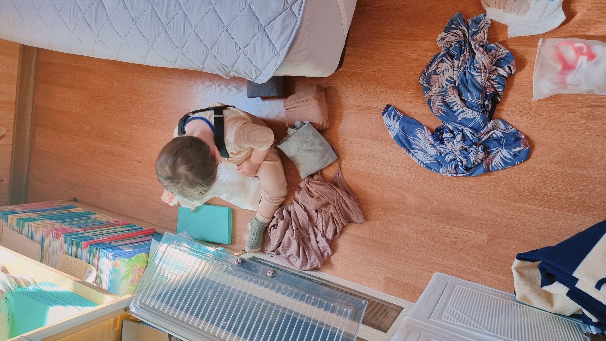 The image, taken from above, shows a child in Dunnae-myeon, South Korea, sitting on a wooden floor next to a bed and a bookcase. The child wears light-colored clothing with suspenders and green socks. Scattered on the floor near the child are folded and unfolded clothes in various colors, including gray, brown, and blue patterned fabric. Other objects visible include diapers, a plastic bag, and clear plastic storage containers. The room is filled with natural light.