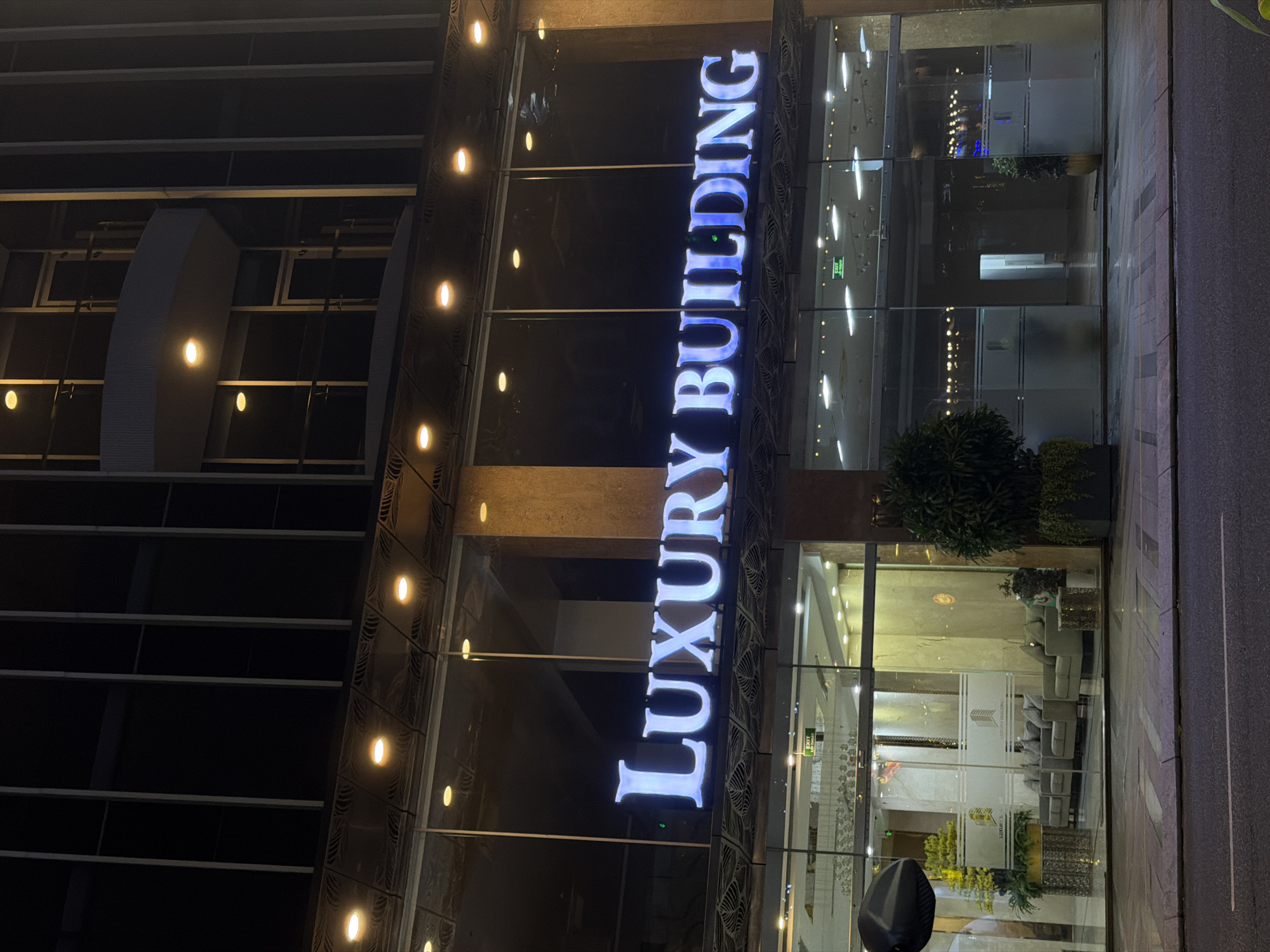 The image shows the exterior of a building at night, with a prominent illuminated sign that reads "LUXURY BUILDING". The building appears to be a modern high-rise with a glass facade. Inside, through the glass doors, a reception area with seating and plants is visible. The setting is likely urban, given the presence of a road in the foreground. The scene is well-lit by artificial lights, both from within the building and from external lighting fixtures. The overall impression is one of an upscale commercial or residential establishment. This building is located on Tran Hung Dao Street in the Son Tra District of Đà Nẵng, Vietnam.