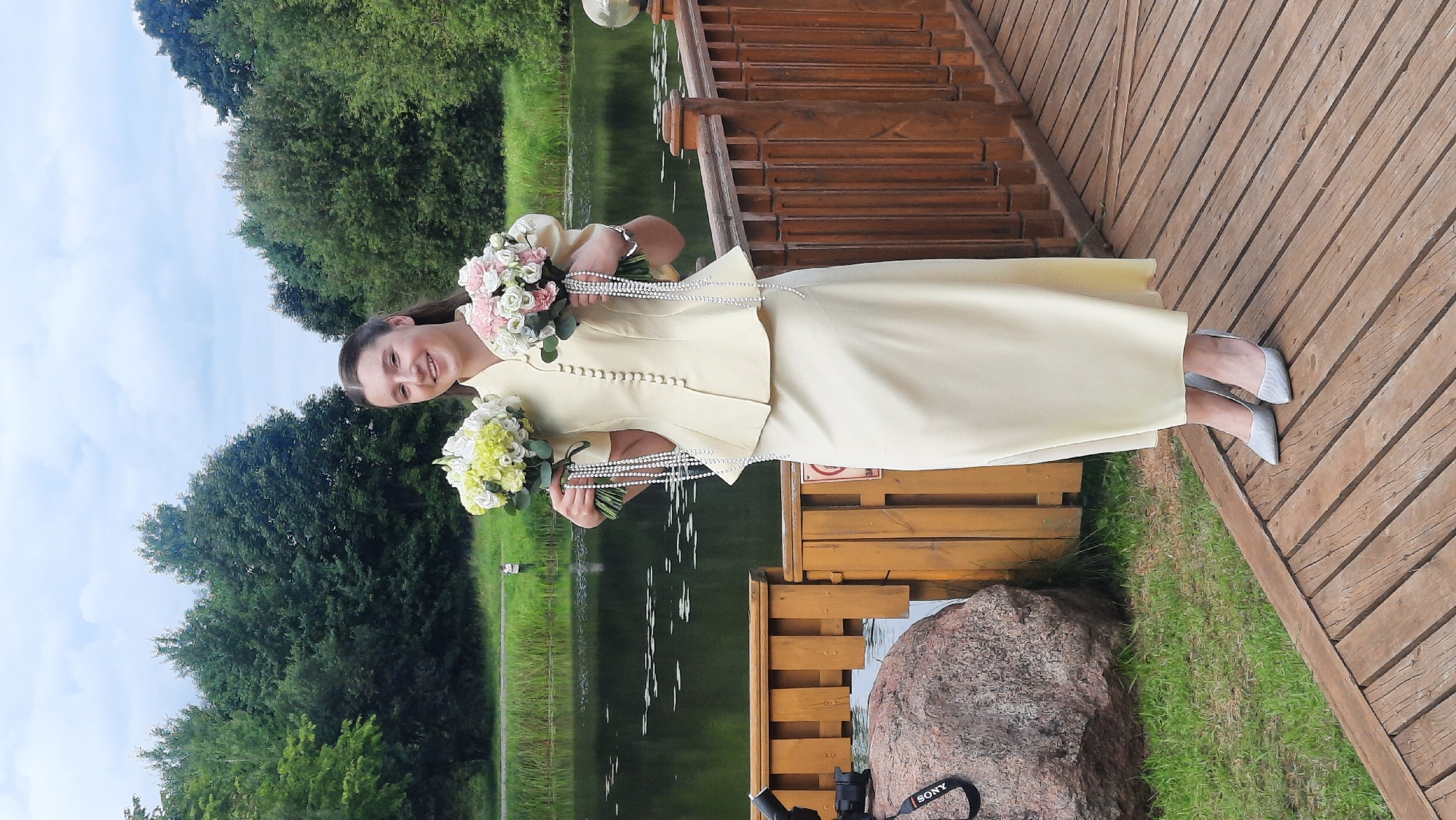 A young woman is standing on a wooden boardwalk, holding two bouquets of flowers. She is wearing a light yellow, peplum-style jacket with a row of buttons down the front and a matching floor-length skirt. Her hair is pulled back, and she is smiling. One bouquet contains white and pale green flowers, and the other has pink and white flowers. Both bouquets are adorned with strands of white beads. Behind her is a body of water, likely a lake or pond, with trees lining the far bank. The boardwalk appears to be part of a park or natural area. The weather is overcast. The location is identified as Жодзішкаўскі сельскі Савет, Belarus.
