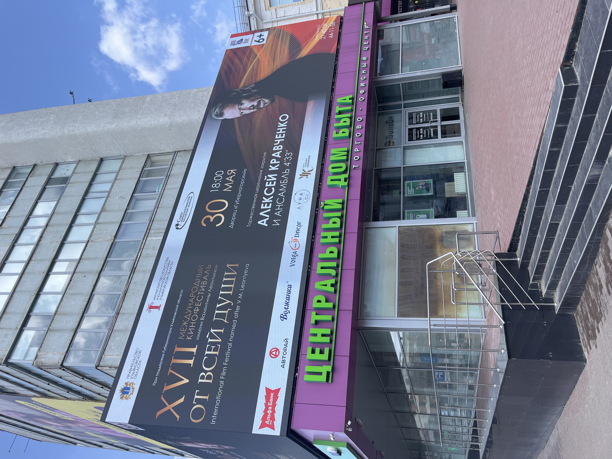 The image shows the entrance to the "Central House of Life" shopping and office center in Ulyanovsk, Russia. A large banner promoting the "XVII International Film Festival 'From the Heart'" is displayed on the building's facade. The banner features a portrait of actor Alexei Kravchenko and advertises a closing ceremony at the Governor's Palace on May 30th at 18:00 featuring Kravchenko and the ensemble 4'33". The banner includes logos of sponsors such as Alfa Bank, Volga Dnepr, and the Luna Film Foundation. The entrance to the shopping center has glass doors and windows, and the name of the building "Central House of Life" is prominently displayed in green letters on a purple background. A staircase with metal railings leads to the entrance. A "Beeline" store is located within the building.