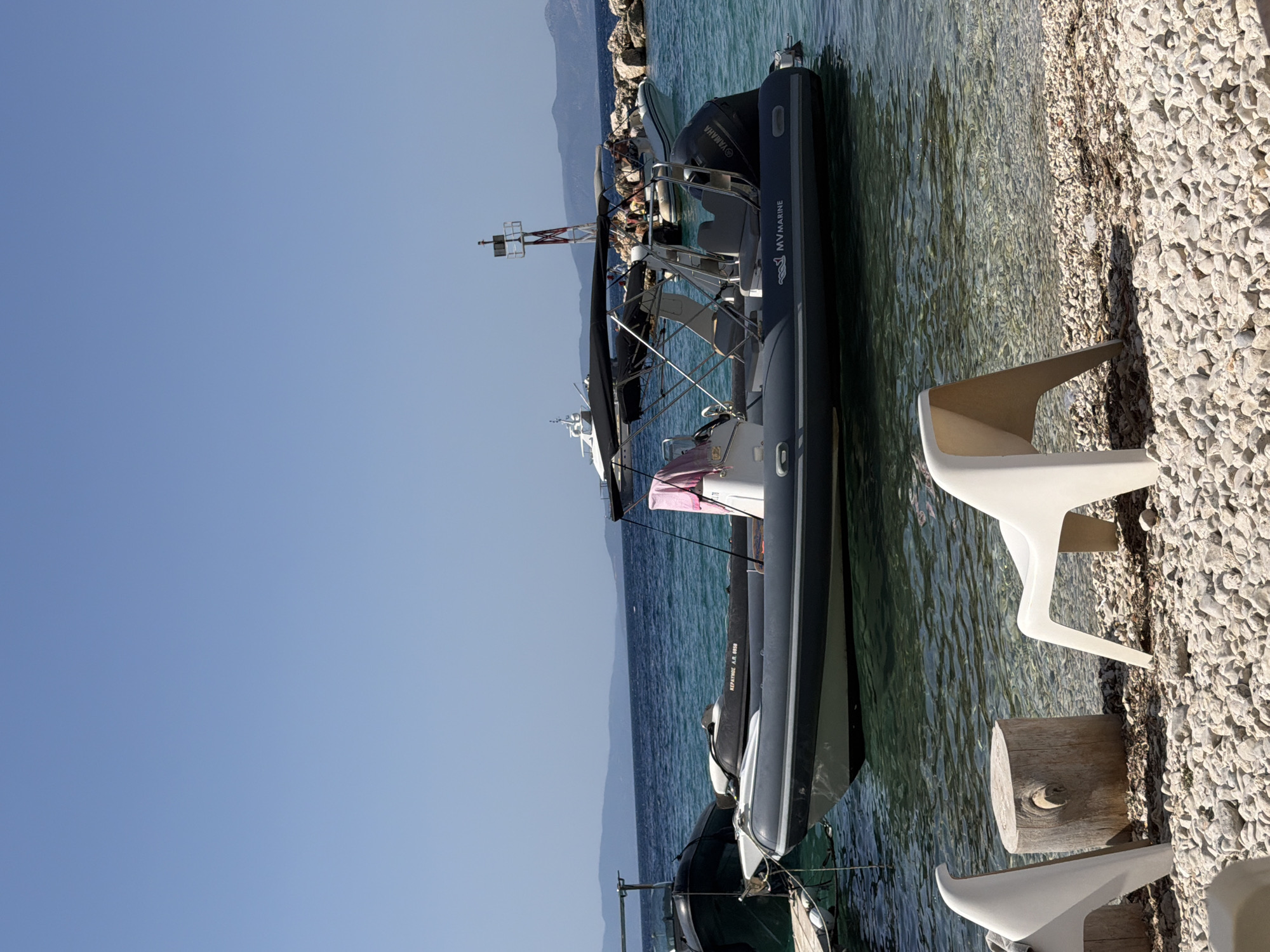 In the Municipal Unit of Kalamos, Greece, a tranquil scene unfolds on a pebble beach with clear, turquoise water. Several inflatable boats are moored in the shallow water. One prominent gray and white inflatable boat, identified by the "MYMARINE" logo and a "YAMAHA" outboard motor, is positioned centrally, showcasing its canopy and a pink towel draped over a seat. To its left, another dark inflatable boat is partially visible, tethered to a wooden dock. Further in the background, a white yacht is anchored, and a red and white navigation buoy stands tall in the water. The shore is lined with smooth, light-colored pebbles, with two white plastic chairs and a thick, weathered wooden log serving as a small table. The horizon is marked by a distant, hazy mountain range under a clear blue sky. The overall impression is one of serene coastal relaxation.