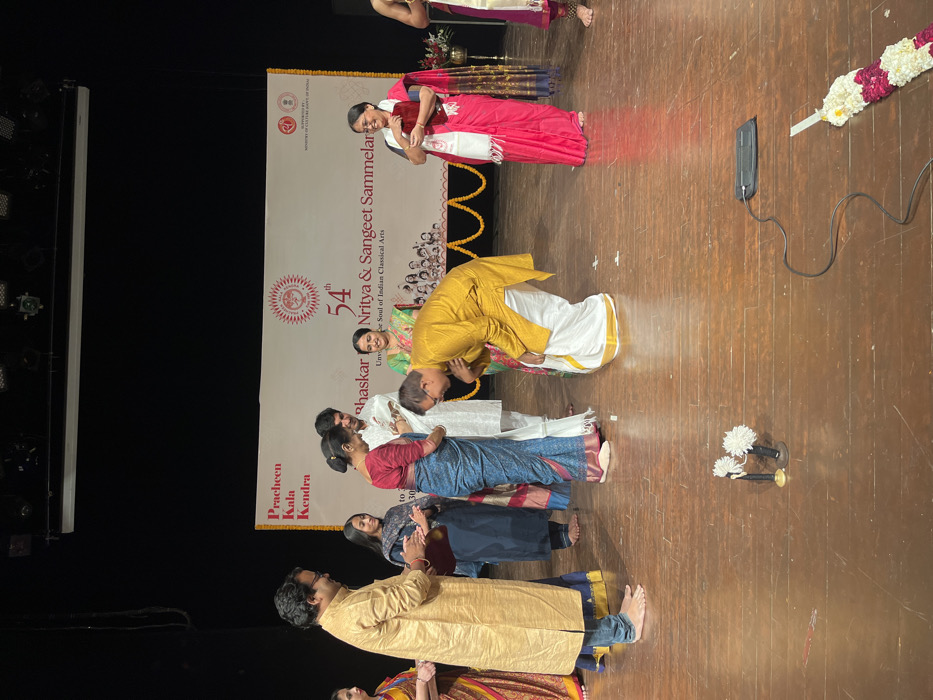 The image shows a group of people on a stage. The stage is made of wood and is decorated with garlands of white flowers. There are two microphones on stands in the center of the stage. The people are dressed in traditional Indian clothing. A man in a yellow and white outfit is bowing to a woman in a red and white sari. There are several other people in the background, including a woman in a blue sari, a woman in a red sari, and a man in a yellow outfit. The stage is decorated with a banner that reads "54th Bhaskar Nritya & Sangeet Sammelan". This event is taking place at Pracheen Kala Kendra in Chandigarh, India (latitude: 30.733, longitude: 76.789).