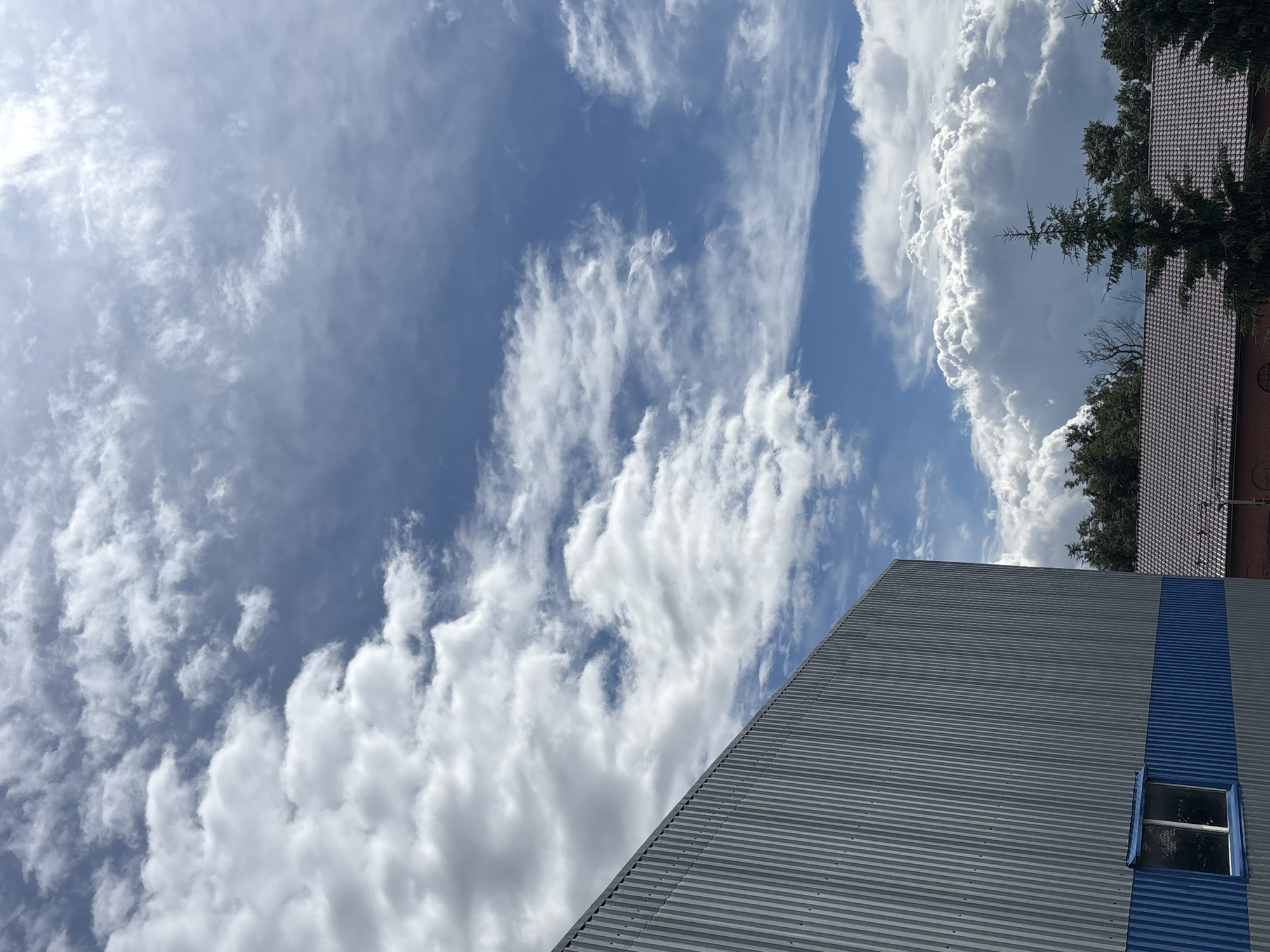 The image captures a daytime exterior scene in Ostrov, Russia, featuring a large, corrugated metal building with a blue stripe near the bottom and a single window. The building's side is angled towards the right. In the background, trees and a tiled roof structure are visible. The sky is filled with a mix of fluffy white clouds and lighter, wispy clouds against a bright blue sky.