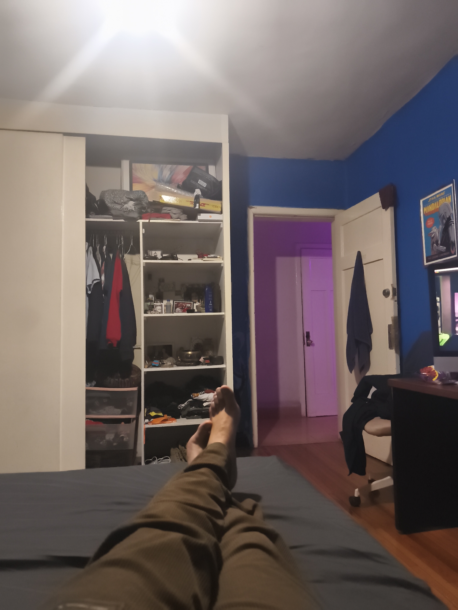 The scene is an interior bedroom setting. The viewpoint is from someone lying on a bed, looking towards a wall with a built-in wardrobe and a doorway. The foreground is dominated by the person's legs and feet, clad in brown trousers, extending towards the wardrobe.
The wardrobe features white sliding doors on one side and open shelving on the other, containing clothes, storage containers, and various personal items. Beyond the wardrobe is a wall painted two shades of blue.
A doorway is visible on the right side of the image, leading to an adjacent room illuminated with a purple glow, where another white door is visible. To the right of the doorway is a desk, an office chair, and a poster of "The Mandalorian" on the wall.
There is no information to indicate the location is in Mexico City, Mexico.