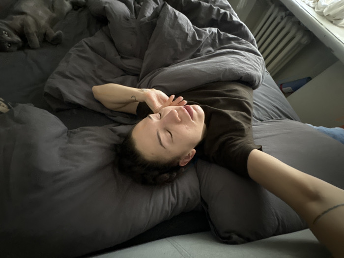 An indoor scene depicts a bedroom in Moscow, Russia. A person, appearing female, lies horizontally on their back in a bed with dark grey bedding, including a fitted sheet, duvet, and pillow. Their head rests on the pillow, with eyes closed or partially shut, suggesting rest or a state of awakening. They wear a dark, short-sleeved top. The person's right arm is bent at the elbow, with the hand near the face, revealing a small crescent moon tattoo on the inner forearm. The left arm is extended towards the bottom right of the frame, exposing a dark band tattoo on the wrist. In the upper left background, a dark grey cat is visible, lying on its back with its head turned towards the viewer. To the right of the bed, a white window frame and a white, ribbed metal radiator are present. Beyond the window, an indistinct urban exterior is partially visible. A blue object, possibly fabric, and other unidentifiable items are discernable on a surface adjacent to the radiator.