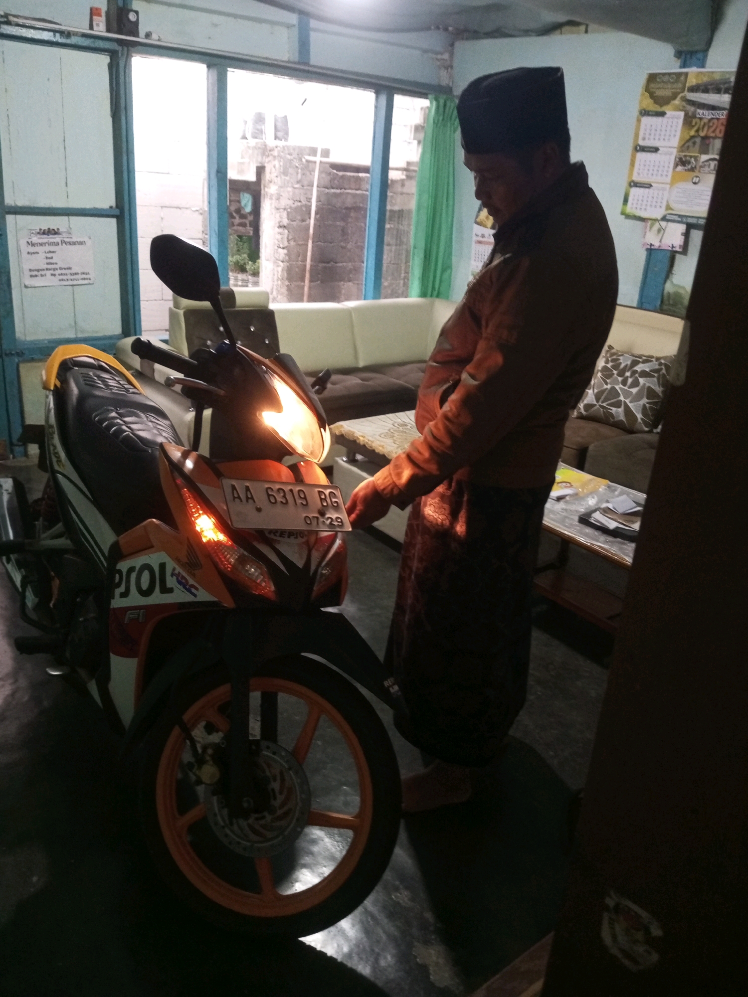 The image depicts an indoor scene, likely a room in a residential property in Andongsili, Indonesia. A man wearing a black peci (cap) and an orange jacket over a patterned sarong stands beside a motorcycle. The motorcycle, an orange and white sports bike with "REPSOL" branding and license plate "AA 6319 BG", is parked indoors, partially illuminated. In the background, a couch and a coffee table are visible, with a calendar from 2026 hanging on the wall. A window on the left reveals a brick wall and some vegetation outdoors. The overall lighting is dim, suggesting it might be evening or an overcast day.