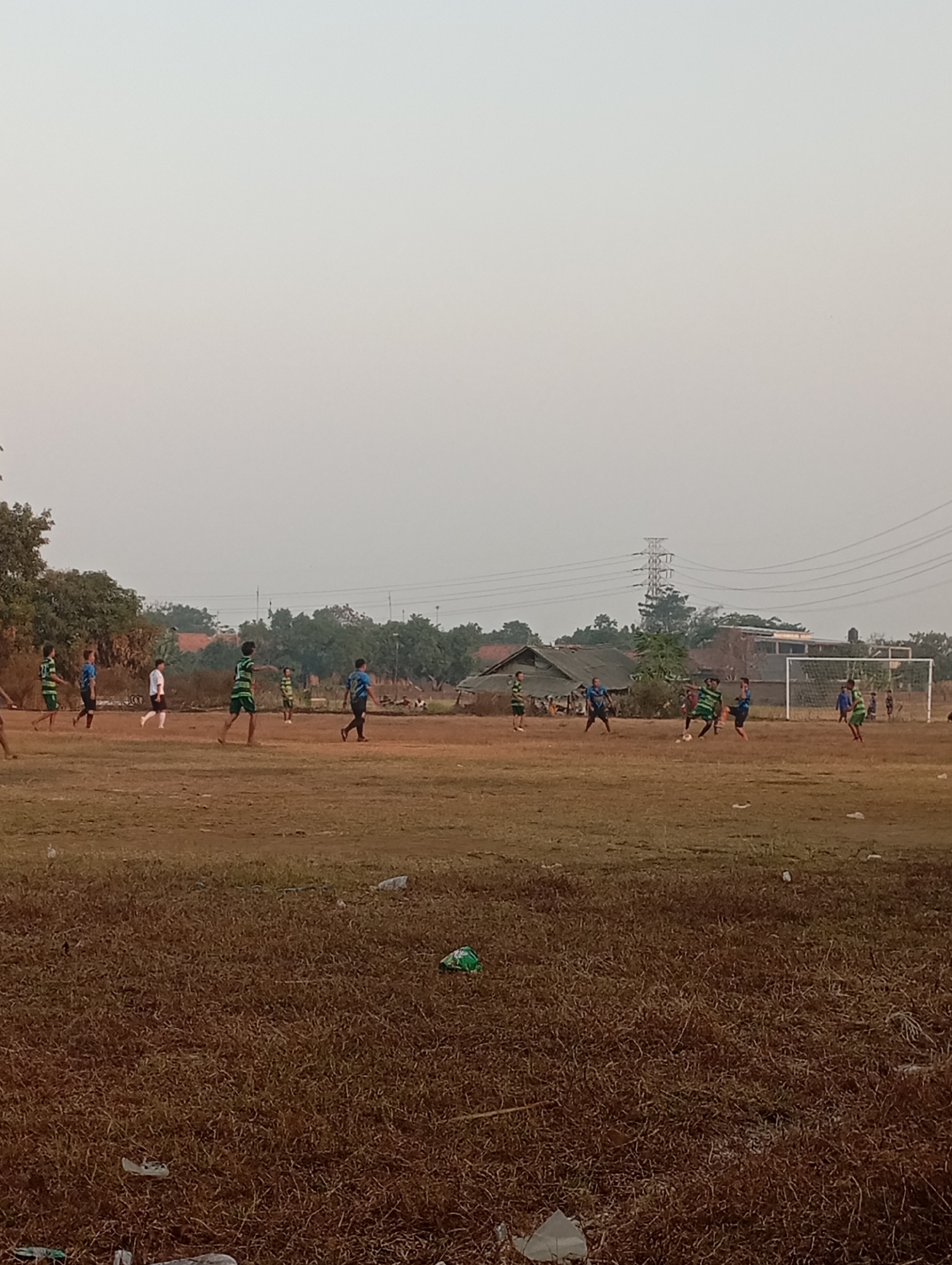 In Kecamatan Haurgeulis, Indonesia, a group of young men are playing soccer on a dirt field. The sky is overcast and hazy. Trees and a few buildings are visible in the background. There is a soccer goal in the distance on the right. The field is uneven and covered with dry grass and debris, including plastic bags. Some of the players are wearing green and white striped jerseys, while others are wearing blue jerseys. The players are engaged in a game, with some actively playing the ball and others observing. The overall scene depicts a casual, informal soccer match in an outdoor, rural setting.