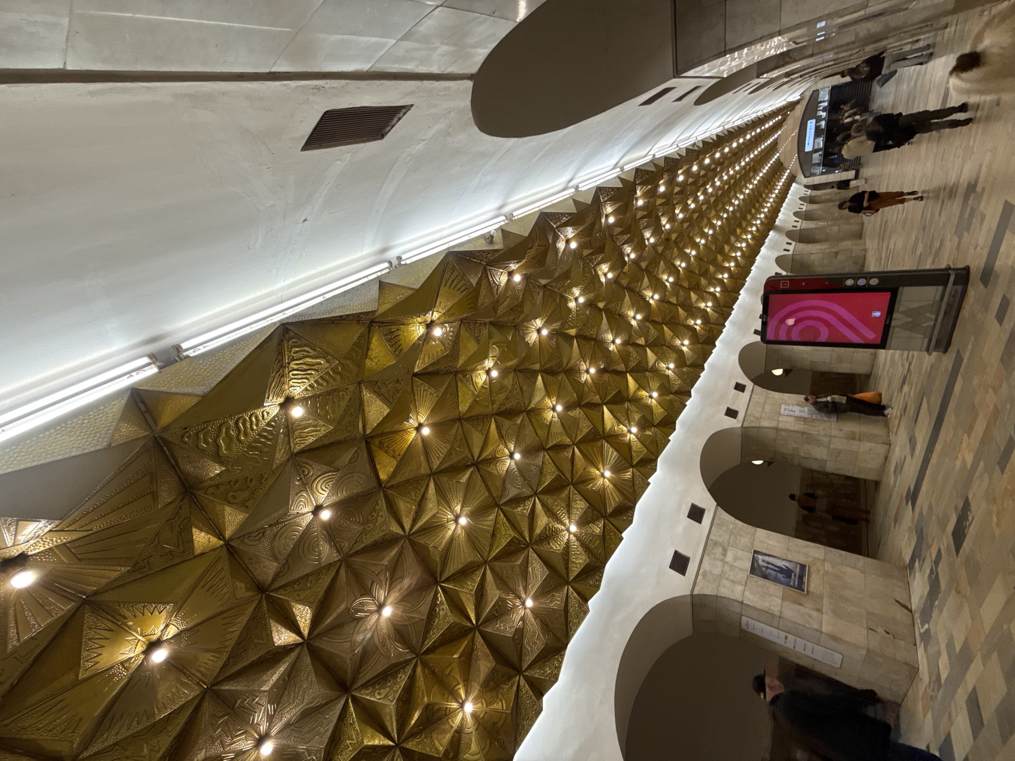 The image shows the interior of a Moscow Metro station, characterized by its ornate golden vaulted ceiling adorned with intricate geometric patterns and illuminated by numerous recessed lights. The ceiling curves downwards, meeting a lighter, more modern section of the ceiling that runs along the length of the platform, interspersed with long, fluorescent light fixtures. The walls of the station are made of beige stone, with large arched openings that lead to presumably other parts of the station or access points. The floor is paved with a mosaic of rectangular and square tiles in shades of brown, grey, and beige. Several people are visible on the platform, walking in various directions. Some are standing, while others are in motion, suggesting a typical day at a busy metro station. One person in the foreground on the left is wearing a dark jacket and a baseball cap, looking away from the camera. Further down the platform, a digital advertising display with a bright pink graphic stands prominently. The overall impression is one of grandeur and historical significance, blended with modern functionality.