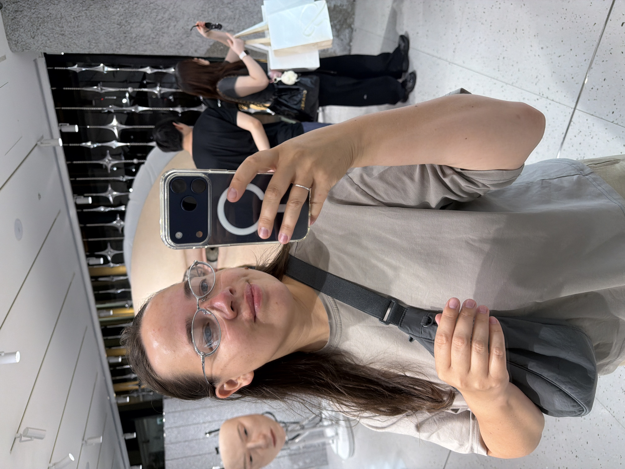 The image captures a woman taking a selfie in what appears to be a well-lit indoor exhibition or retail space, possibly in Chuo, Japan. She is positioned in the foreground, wearing a light-colored t-shirt and a crossbody bag. Her face is framed by silver-rimmed glasses, and she is holding a smartphone to capture the photo. In the background, several art installations or displays are visible, including a manikin head on a stand to the left and a large, abstract sculpture or display behind her. Further in the background, two other individuals are seen, one man in a black t-shirt and a woman holding shopping bags. The environment features clean, modern architecture with white ceilings, speckled floors, and metallic decorative elements on a dark wall. The lighting is bright and even, suggesting an indoor commercial or public space.