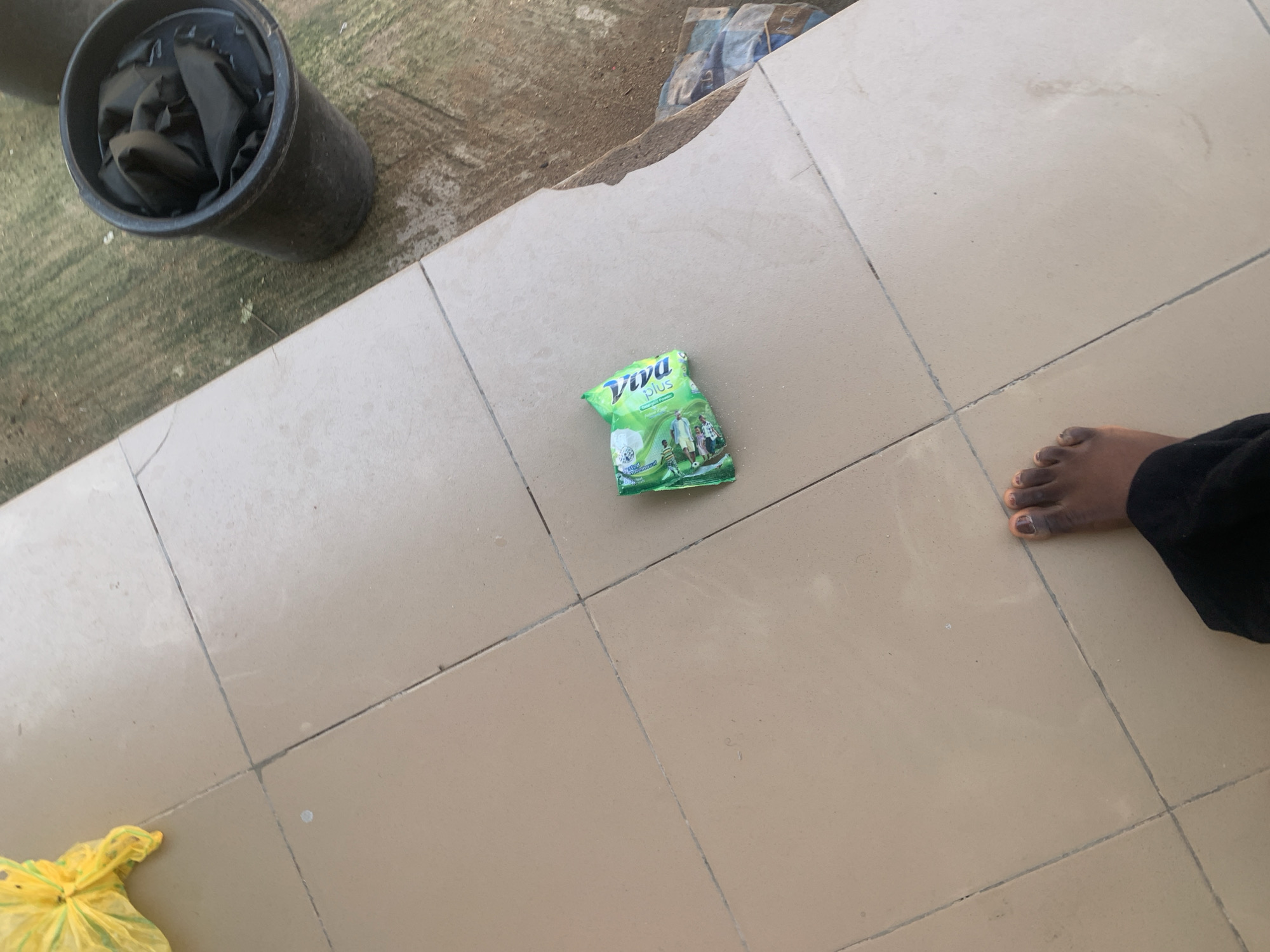 In Jauro Yinu, Nigeria, an outdoor scene is depicted on a tiled surface. A yellow plastic bag, partially visible in the upper left, lies near the edge of the frame. In the center, a small green packet of "Viva Plus" detergent is placed on the tiles. To the right of the detergent packet, a dark-colored bucket filled with black fabric is situated on a rough, mossy ground. In the lower portion of the image, a person's bare foot with painted toenails is visible, wearing dark trousers. The overall environment suggests a utilitarian or residential outdoor space.