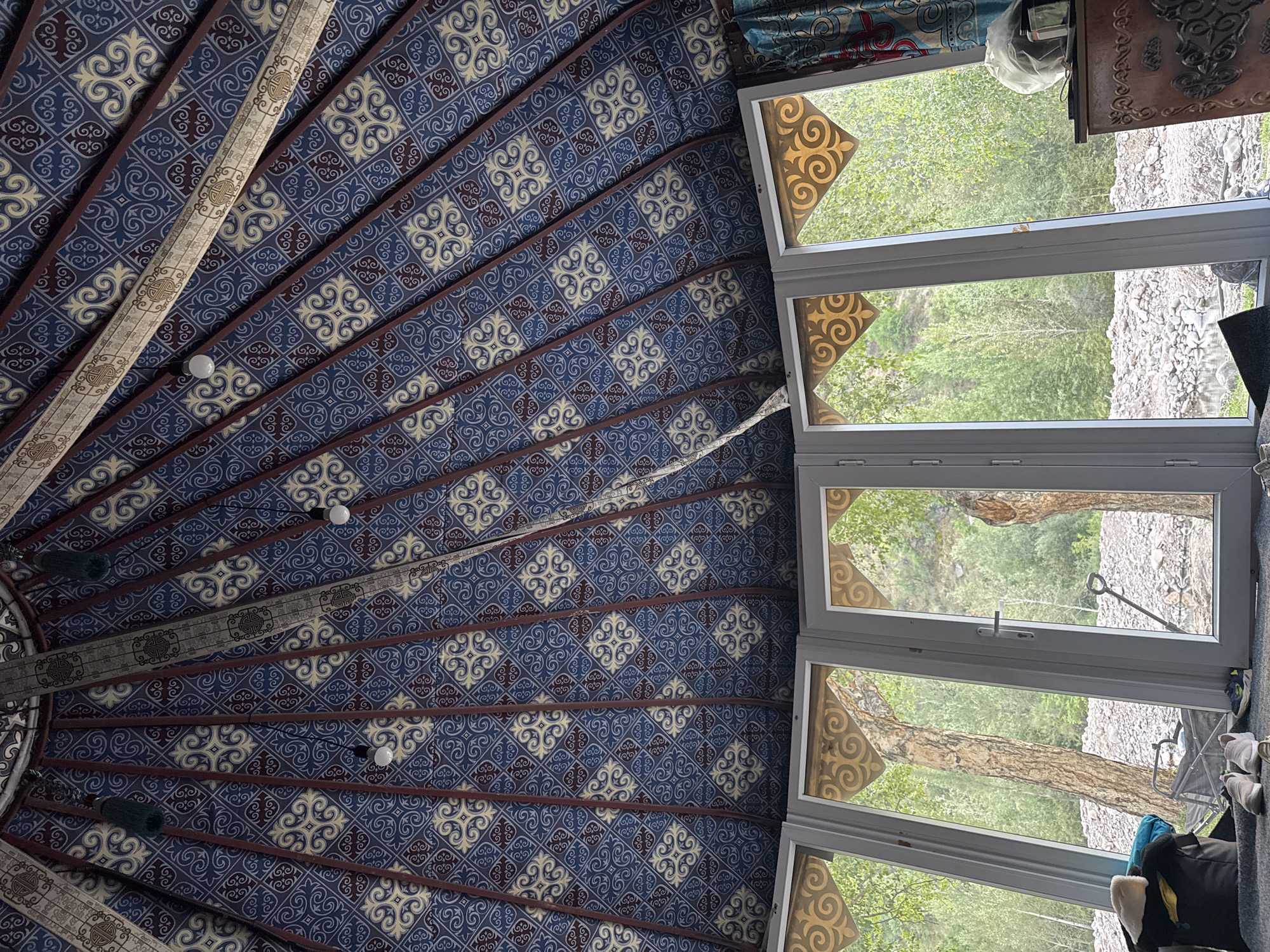 The image captures the interior of a yurt, showcasing a decorative ceiling with a blue and yellow patterned fabric supported by dark brown beams. Three lamps hang from the ceiling. A set of large, white-framed windows dominates the lower part of the image, revealing a lush, green outdoor environment. Outside, trees, rocky terrain, and a small body of water are visible. In the foreground inside the yurt, near the windows, are a few personal items including a black bag, a pair of shoes, and some folded clothing. A small, brown, ornate chest or cabinet is partially visible on the right side of the frame. The scene appears to be in a natural, possibly remote location, consistent with Leskhoz, Kazakhstan, given the yurt structure and surrounding landscape. There are no people visible in the frame.