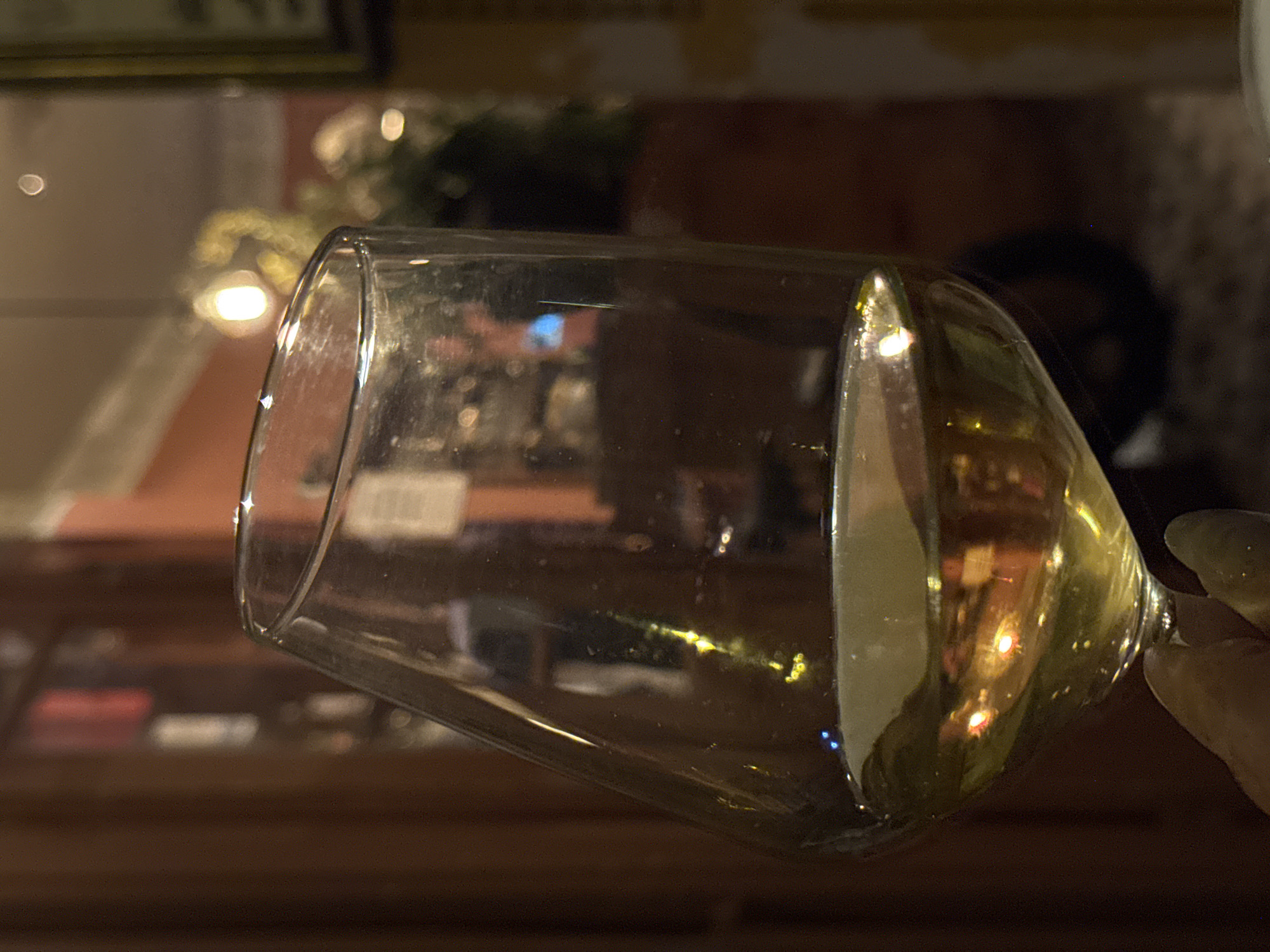 A close-up, low-angle shot focuses on a tilted wine glass partially filled with a yellow liquid, likely white wine. The glass appears to be held by a person's hand, visible on the right side of the frame. The background is softly blurred but appears to be an interior setting, possibly a restaurant or bar, with warm lighting. Reflected in the glass are shapes and light, suggesting other patrons and ambient lighting. No specific details or landmarks indicate the exact location as Tel-Aviv, Israel.