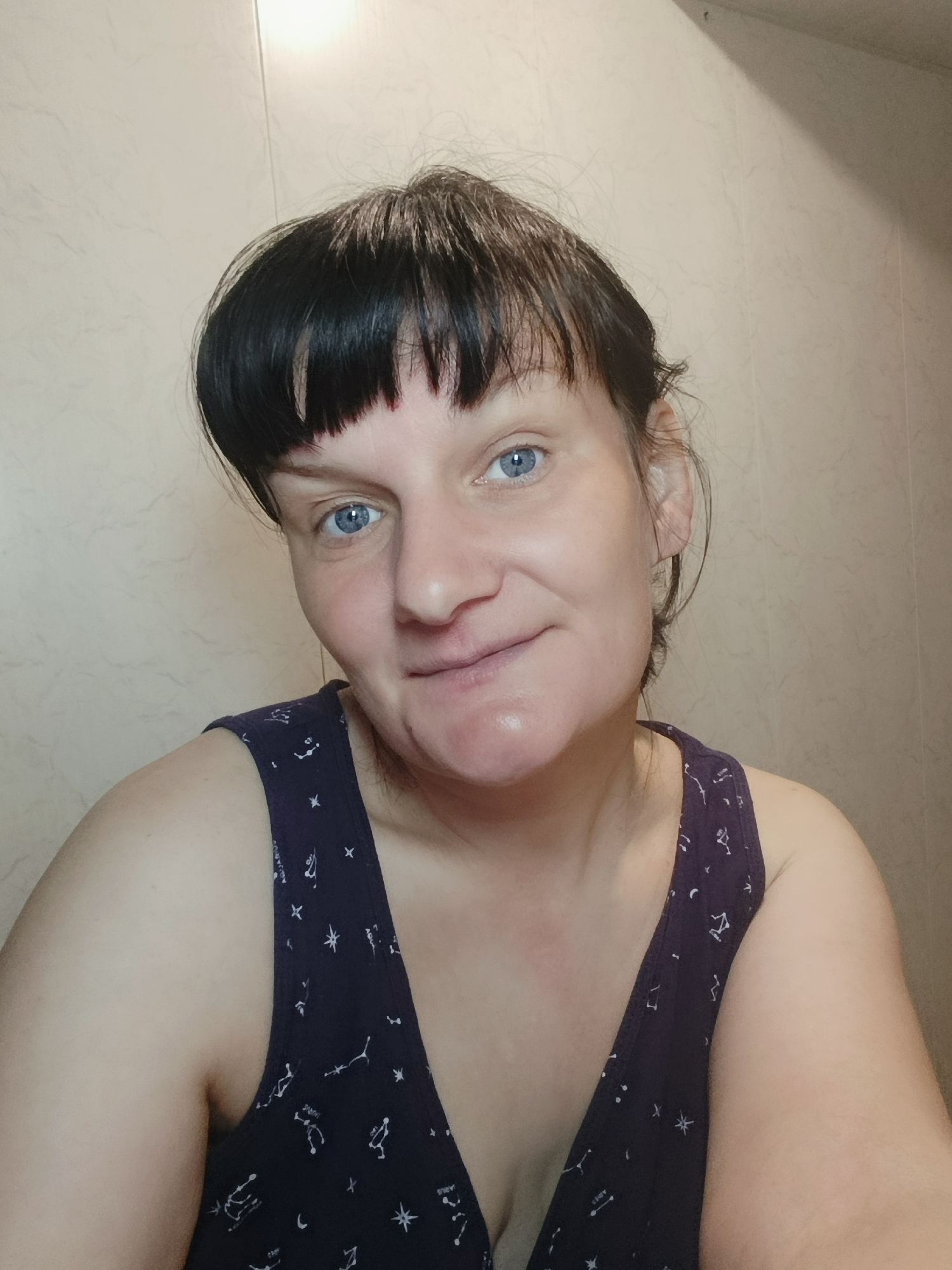 A woman with short, dark hair and blue eyes is centered in a close-up, chest-up shot. She is wearing a dark blue tank top patterned with white constellations. The background is a light beige wall with subtle vertical lines, possibly paneling. The lighting is bright and even, casting minimal shadows. The woman has a slight smile. There is no evidence to confirm the location as Фоки, Russia.