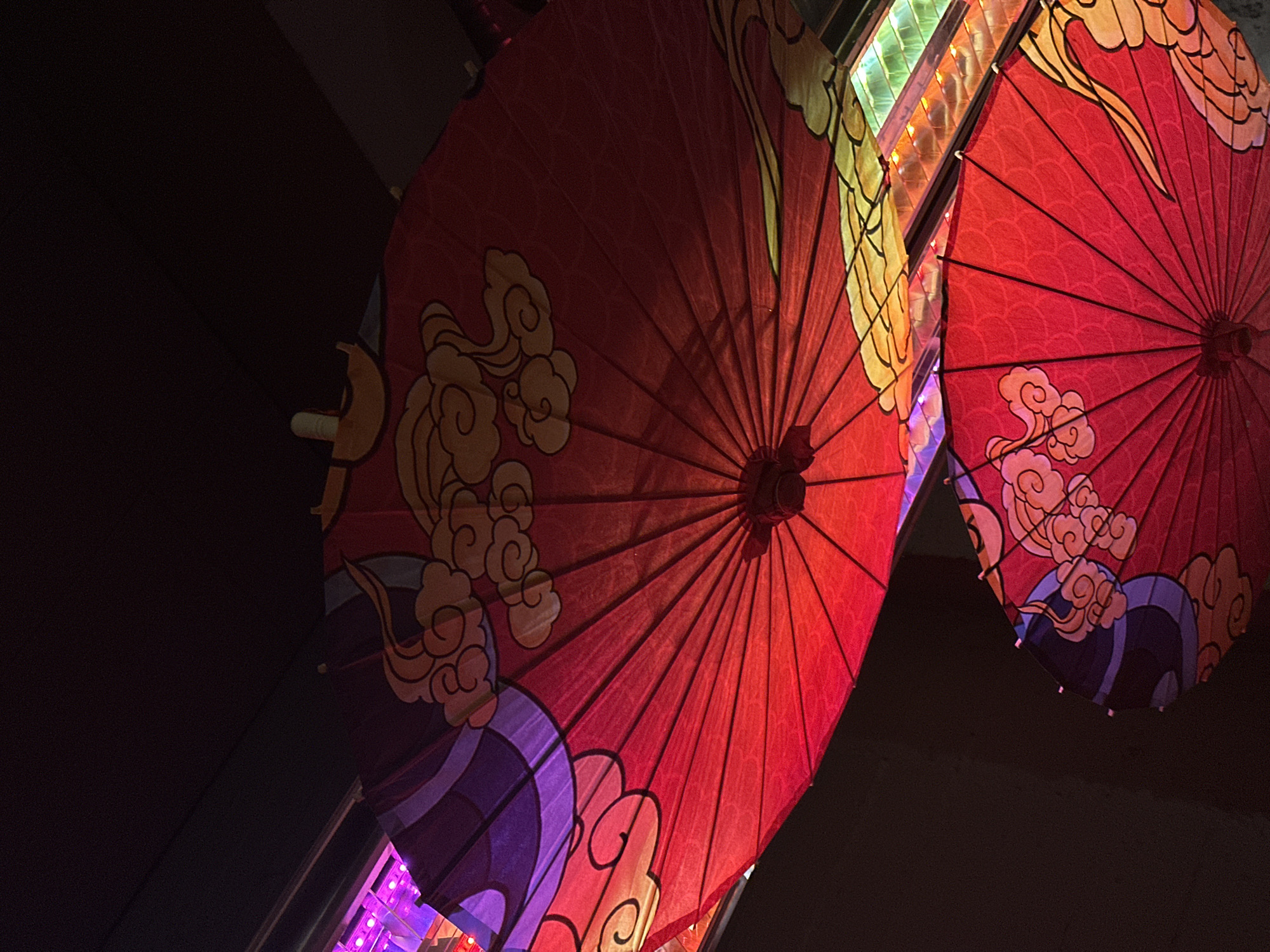 Two red, open umbrellas with decorative designs of clouds and purple waves are hanging from above. The umbrellas are positioned at an angle, with the top umbrella larger and more prominently displayed. The umbrella designs are intricate, featuring swirling cloud patterns in yellow and orange hues, as well as bold purple wavy lines on the upper umbrella. The background is dark, with hints of colorful lights, possibly from a ceiling or decorative lighting, in shades of pink and green visible between the umbrellas. The image has a slightly tilted perspective, as if captured from below. The location is not identifiable as Paris, France.