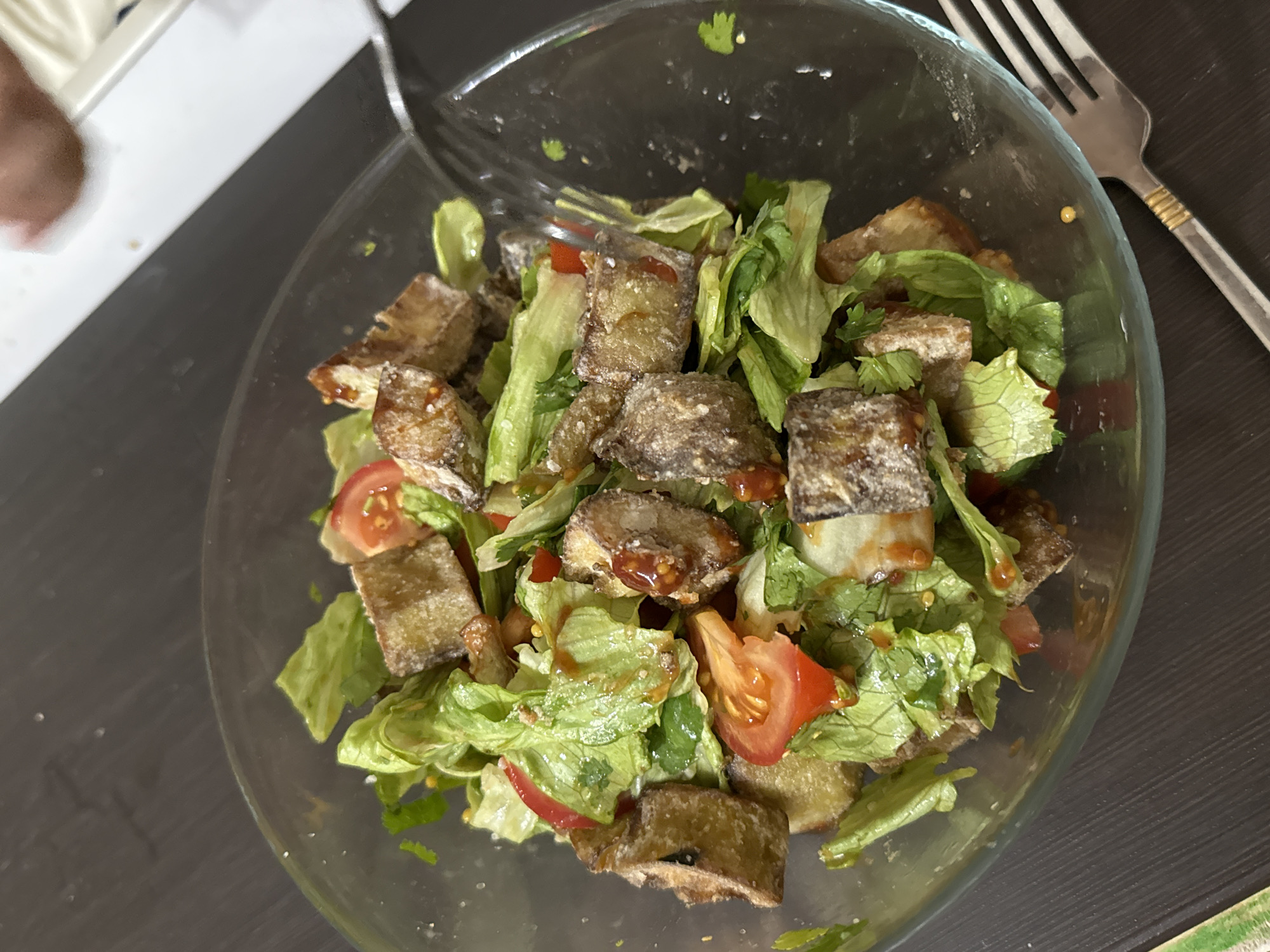 A close-up, high-angle shot captures a freshly prepared salad served in a clear glass bowl, positioned on a dark, possibly black or dark gray, surface. In the upper left background, a lighter, off-white surface is partially visible, along with a blurred brown shape that could be a person's arm or hand.
The salad consists primarily of roughly chopped green lettuce as its base. Scattered throughout are several chunks of what appear to be fried or baked eggplant, or possibly tofu, characterized by their light brown, somewhat textured, and slightly crispy exteriors. Some of these pieces have a reddish-orange dressing drizzled over them. Bright red tomato wedges or slices are interspersed among the greens and main protein/vegetable, adding a pop of color. Small sprigs of green herbs, possibly cilantro or parsley, are also mixed into the salad.
A silver fork with a distinctive gold-colored band on its handle rests partly within the bowl, suggesting the salad is either about to be eaten or is currently being consumed. The lighting is consistent with an indoor setting, but the time of day is not discernible. There is no visible text in the image. The scene depicts a simple meal being presented. No specific location cues are present within the image itself to indicate Chaikovsky, Russia, other than the provided context.