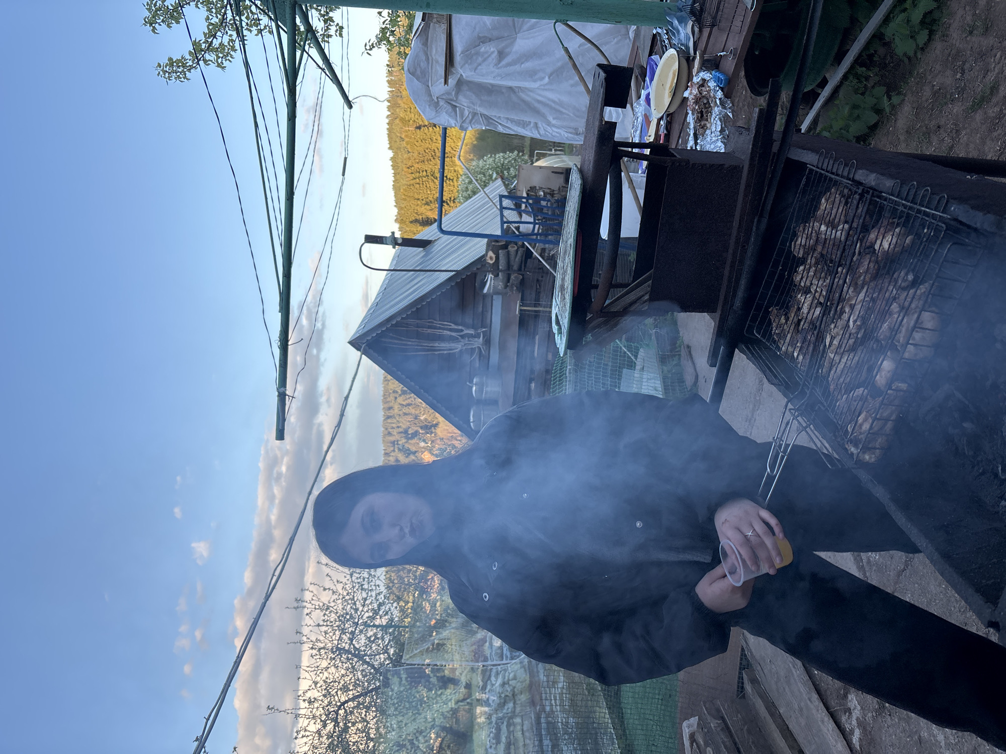 A person is grilling meat in Фоки, Russia. The person is standing next to a barbecue grill with a wire grate containing pieces of meat. The grill is surrounded by smoke. The person is wearing a dark hooded jacket and holding a clear plastic cup with a yellow liquid inside. In the background, there is a wooden building with a metal roof, trees, and a blue sky. There is also a covered structure and other objects on a deck or patio area.