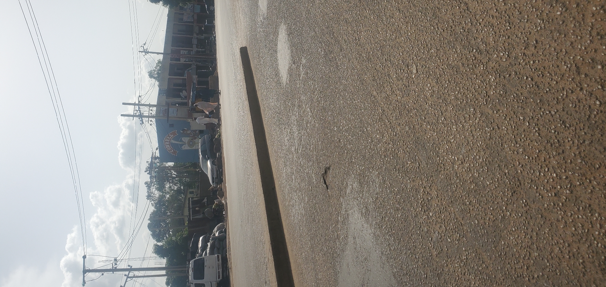This image depicts a street scene in Jos, Nigeria, on a bright, overcast day. The foreground is dominated by a wet asphalt road with visible puddles and a curb. In the mid-ground, a white van is parked on the left side of the road, next to a pile of large sacks, possibly containing goods. To the right of the van, a silver station wagon is parked. Further to the right, a building with a blue mural that says "COWBELL" is visible. Three individuals are standing near the building: two men wearing traditional white attire and a woman in a light-colored garment. The background features utility poles and trees under a cloudy sky. The overall impression is a bustling street scene with commercial activity and everyday life in Jos.