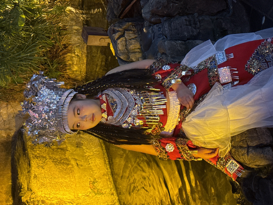 The image depicts a young individual, identified as a child, seated and facing slightly left in an ornate, traditional-style costume. The scene is located in Yuzhong District, China. The individual wears a large, elaborate silver-toned headdress adorned with intricate designs and dangling elements, atop long, dark braided hair. A multi-layered silver-toned necklace with numerous pendants covers the chest. The attire includes a red top or vest and a long red skirt, both heavily embellished with silver-colored metallic plaques, intricate embroidery, and dangling ornaments. A white, sheer, ruffled fabric layer is visible over the red skirt. The individual's gaze is directed off-camera to the left, with a neutral expression; hands are clasped or resting in the lap, partially obscured by the skirt.
The subject is positioned on dark, irregular rock formations. To the left, a body of water reflects a warm, yellowish-orange light. The background behind the subject on the left features a textured, yellowish-orange illuminated rock-like wall or structure. To the right and behind, dark green foliage is visible, along with additional dark rock formations and a distinct dark, rectangular object, possibly made of wood or metal, resting on the rocks. The environment is characterized by artificial, warm-toned lighting, creating a somewhat dim atmosphere.