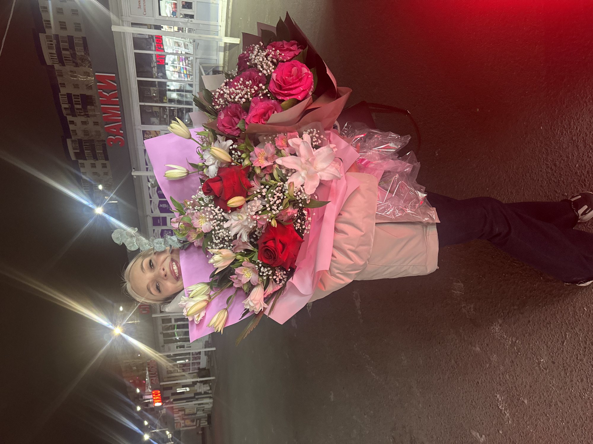 The image shows a woman holding two bouquets of flowers outdoors at night. She is smiling at the camera, with bright lights behind her. The larger bouquet has red and white roses and lilies wrapped in pink paper, while the smaller bouquet contains smaller pink roses wrapped in brown paper. She is wearing a light jacket and dark pants and sneakers. The background reveals a building with the word "ЗАМКИ" lit up in red. The image was taken in Surgut, Russia.