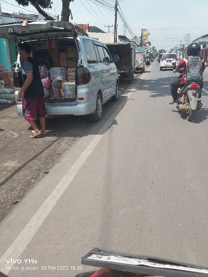 This image depicts a vibrant street scene in Patrol, Indonesia, captured during late morning on a sunny day. In the left foreground, a silver minivan is parked, its rear hatch wide open, revealing a cargo area heavily loaded with various packaged goods, including boxes, bags, and bottled drinks. A man, wearing a black t-shirt, red and black patterned shorts, and flip-flops, stands by the open trunk, appearing to be loading or unloading merchandise.
The paved road stretches from the foreground into the background, marked by a faded white line on the left edge. On the right side of the road, a person on a motorcycle, wearing a black helmet and a grey backpack, is visible, suggesting active traffic. Further down the street, other vehicles, including a white car and a truck, can be seen, indicating a moderately busy thoroughfare. Overhead, a dense network of power lines spans across the sky. Residential and commercial buildings line the street in the distance.
The scene conveys a sense of everyday activity and commerce in a typical Indonesian setting. Visible text includes a phone watermark at the bottom left stating "vivo Y19s", "Akhtar & Arvan", and the date and time "20 Feb 2025, 10.20". A partial view of a license plate holder is visible at the bottom right, showing some indistinct letters.