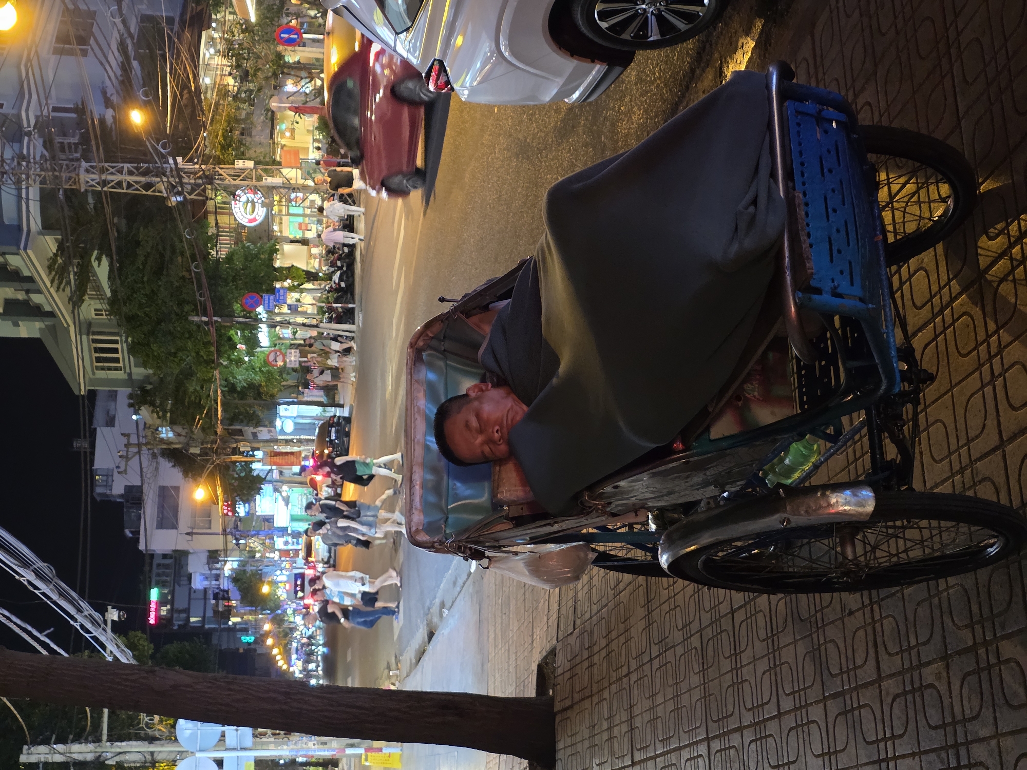 This nighttime image captures a man sleeping peacefully in a blue-framed cyclos (pedicab) parked on a patterned sidewalk in an urban setting. The man is covered with a dark grey blanket, his head resting on a cushion inside the cyclos.
Behind the cyclos, a busy street is visible with cars, including a white and a red vehicle. Numerous pedestrians are walking on the sidewalk and street in the background, indicating an active evening. The street is brightly lit by warm orange streetlights and the colorful, often neon, signs of various shops and businesses lining the multi-story buildings.
Notable details include a dense overhead network of electrical wires, a common sight in many Vietnamese cities. Several circular traffic signs, predominantly red and blue, indicating restrictions like "No Stopping" or "No Parking," are scattered throughout the scene. A tree trunk anchors the left side of the foreground. The bright artificial lighting against the dark sky confirms the time of day is night. The scene conveys a sense of a bustling city continuing its activity late into the evening, with a moment of quiet rest for the cyclos driver. This setting is consistent with Nha Trang, Vietnam.