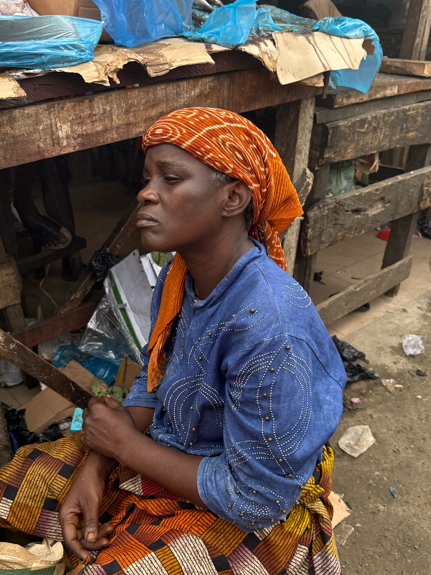A Black woman in a blue denim shirt adorned with white beadwork and an orange and white patterned headscarf sits in a market stall in Lagos, Nigeria. She wears a brightly colored African print skirt with horizontal stripes of orange, yellow, and white, accented by black. Her hands are clasped in her lap, and she is looking to her left with a thoughtful expression. The stall behind her is constructed from rough wooden planks and appears weathered. Various items are visible within the stall, including blue plastic sheeting, cardboard boxes, and what appear to be wrapped goods. The ground in front of the stall is dirt and littered with debris, including plastic bags and wrappers. The overall impression is of a busy, outdoor market environment.