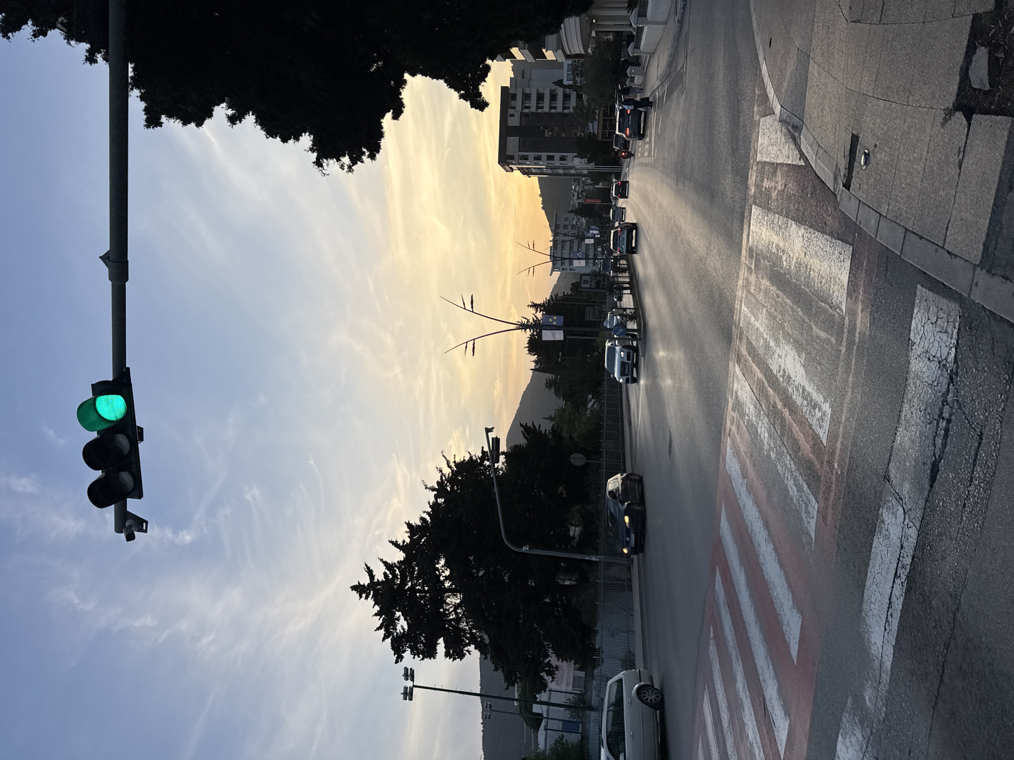 The image shows a street scene in Budva, Montenegro, during sunset. The street is lined with buildings on the right and trees on the left. Cars are driving on the street. A traffic light on the left is showing a green light. A pedestrian crossing is visible on the right side of the street. The sky is a mix of blue and orange due to the sunset. A mountain is visible in the background.