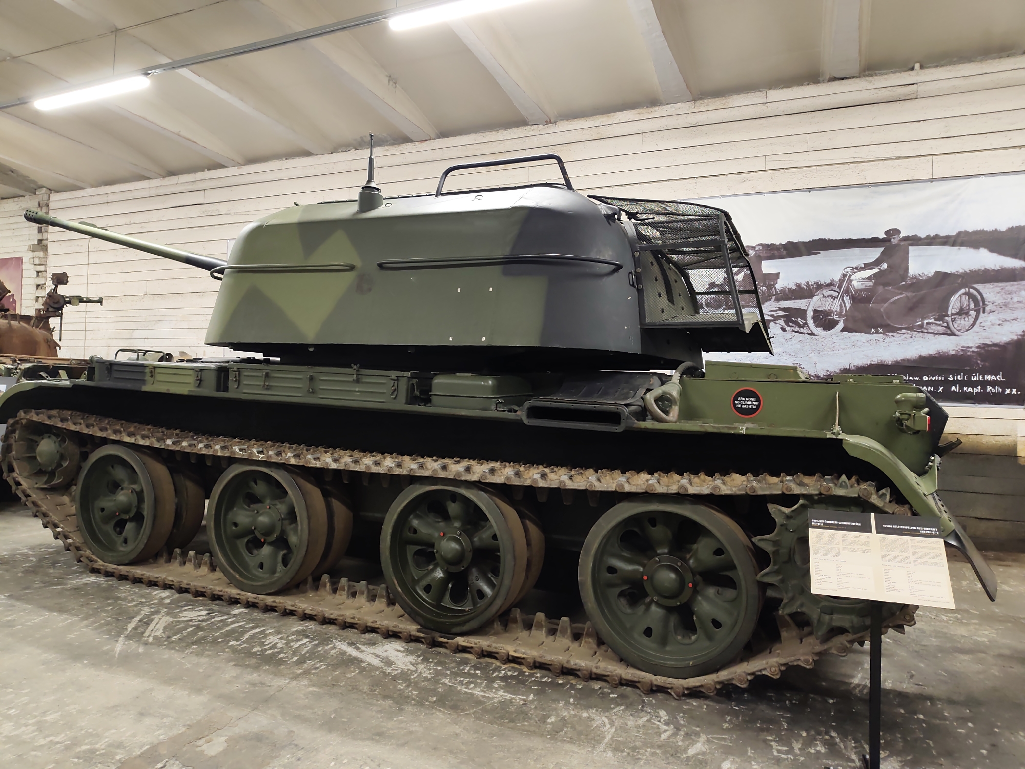 An anti-aircraft vehicle, painted in a camouflage pattern of dark green and light green, is parked indoors on a concrete floor. The vehicle has a long barrel gun extending from the front of the turret. The turret is open on the rear and sides, with a metal mesh cage. The vehicle has a track system with multiple road wheels. To the right of the vehicle, there is a poster on an easel with text and an image of a man on a motorcycle with a sidecar. In the background, there is a wooden paneled wall and fluorescent lighting fixtures on the ceiling.