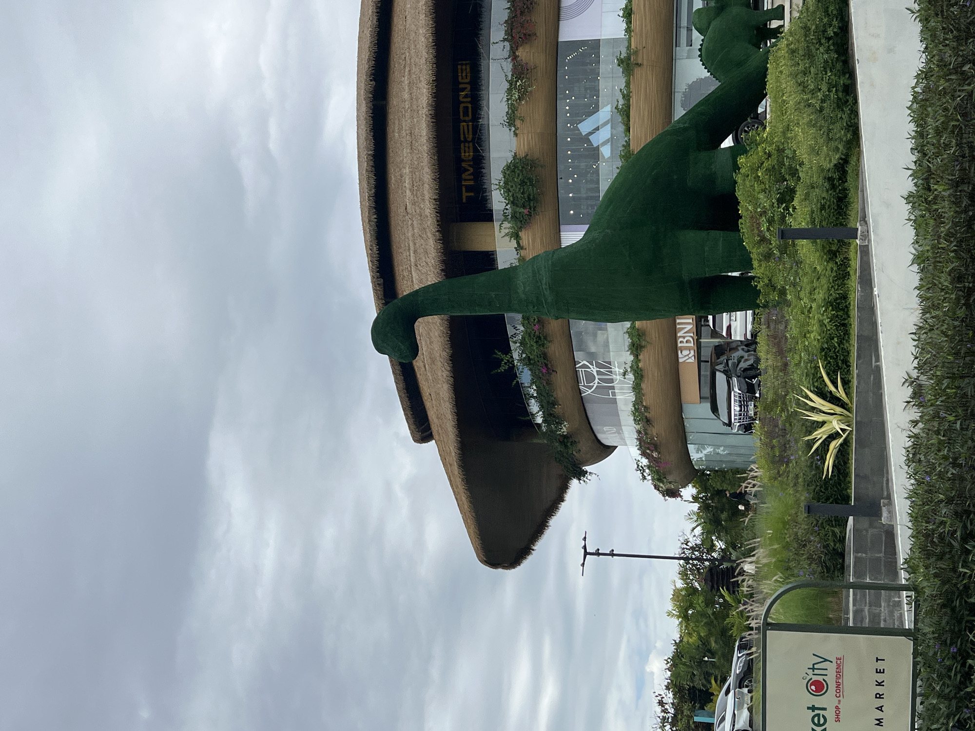 The image shows a large, green, dinosaur-shaped sculpture in front of a modern building with a thatched roof. The building appears to be a commercial establishment, with the text "TIMEZONE" visible on the facade. There are also cars parked in front of the building and a sign that reads "MARKET CITY". The sky is overcast. This scene is located at Level 21 Mall in Denpasar, Bali, Indonesia, which is a well-known shopping destination featuring unique architectural designs and attractions like the dinosaur sculptures.