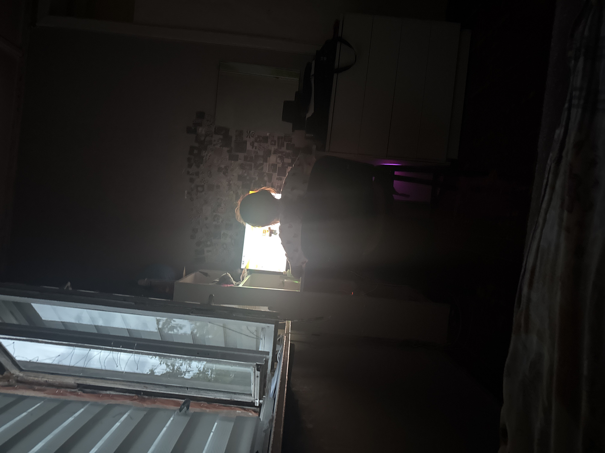 In a dimly lit room, a person is seated at a desk, facing away from the camera, illuminated by the glow of a computer monitor. The room appears to be a bedroom or study, with a bed in the foreground covered in a blanket. To the left, a large window offers a view of the outdoors, with a corrugated metal wall visible on the exterior. On the right side of the room, a dresser with several drawers is present, with a dark bag and a black object placed on top. The wall behind the person is decorated with numerous small pictures or photographs. The lighting suggests it is either early morning or late evening. The environment is consistent with a residential setting in the city of Olkhovka, Russia.