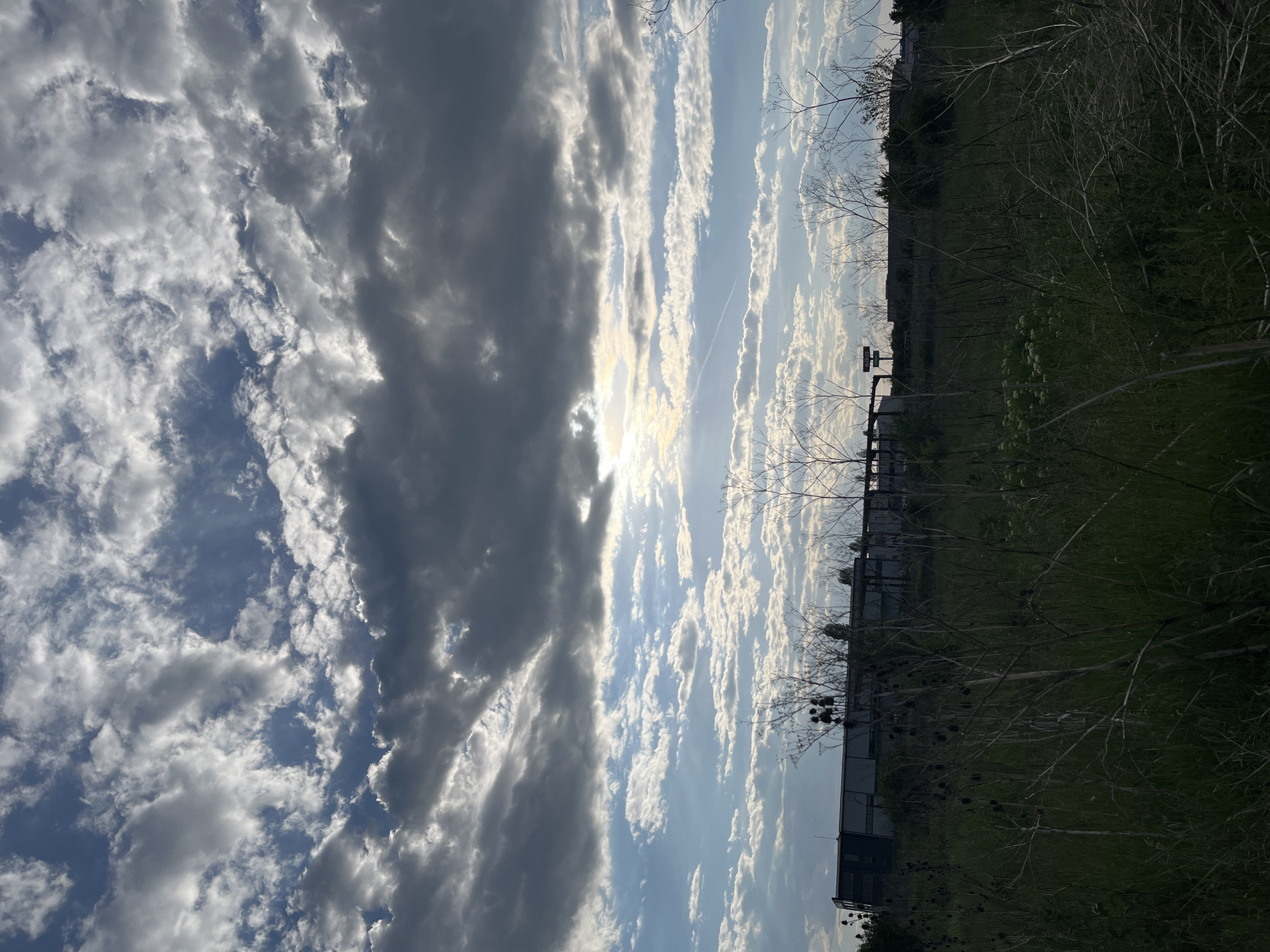The image shows a daytime scene in Sofia, Bulgaria. The sky dominates the upper portion of the frame, featuring a mix of dark, heavy clouds on the left and thinner, brighter clouds on the right, with the sun visible through a gap. A contrail is visible in the sky. The lower portion depicts a grassy area with trees and shrubs, some appearing bare. A fence and a building can be seen in the background. A sign is also visible near the fence.