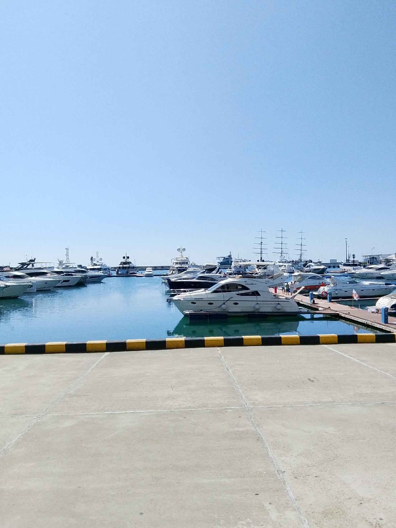 This image shows a marina filled with numerous yachts and boats of various sizes docked in calm blue water under a clear sky. In the foreground, there is a concrete pier with a yellow and black striped barrier along the edge separating it from the water. The marina extends into the background with more boats, some with tall masts, suggesting a large and well-equipped facility. A few people can be seen on the docks, but their activities are not discernible. The overall scene depicts a bright, sunny day at a picturesque waterfront location.