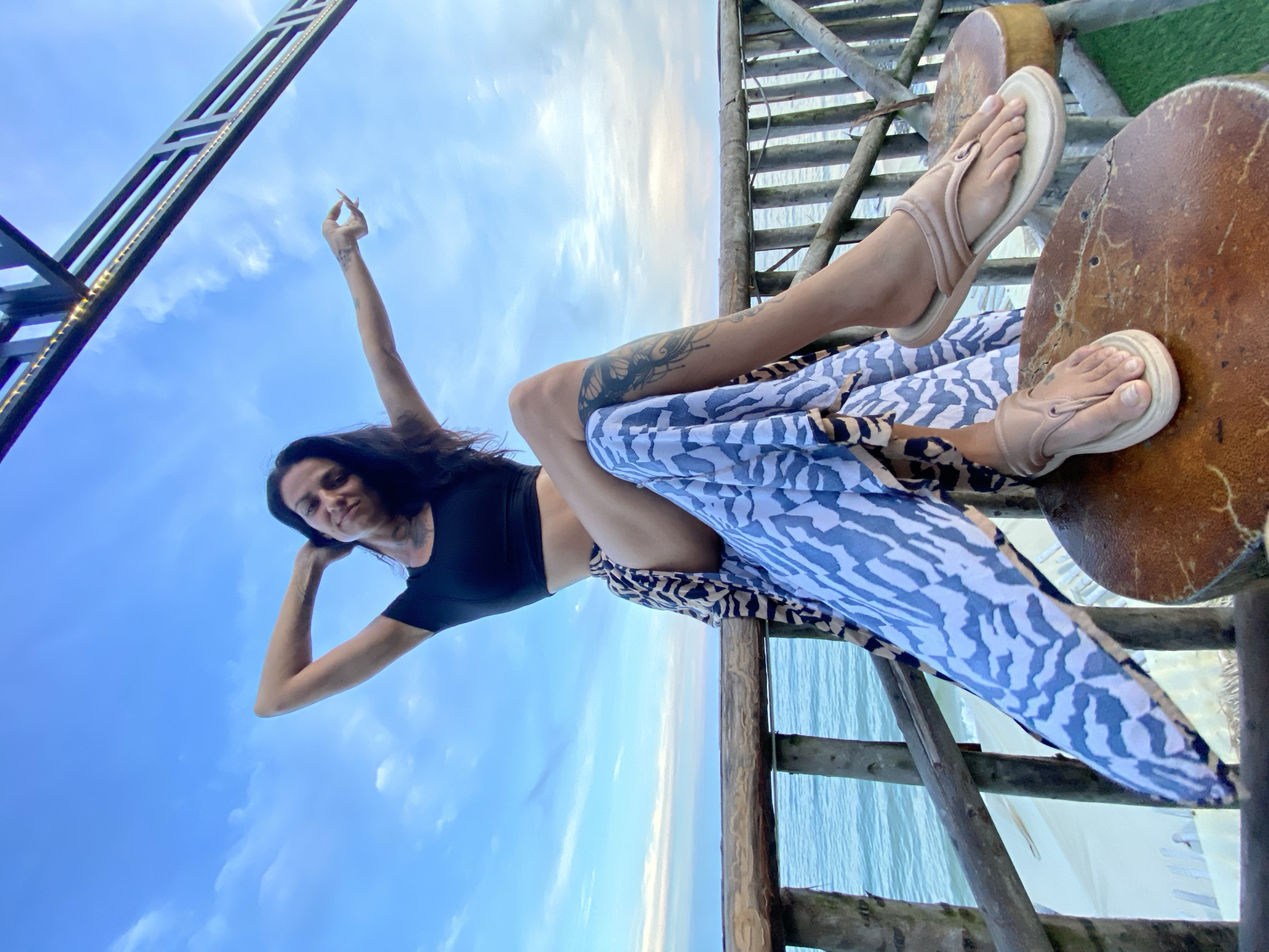 A low-angle, wide shot captures a woman in a black crop top and a patterned skirt posed on a wooden structure overlooking the ocean. She is lying on her side, with her legs propped up on circular wooden surfaces, one foot adorned with a beige sandal. Her arms are raised, and she appears to be holding a cigarette in her right hand. A prominent tattoo is visible on her left leg. The background consists of a vibrant blue sky with scattered clouds, and the ocean is visible below the wooden platform, suggesting an outdoor setting, likely near a beach. The lighting and sky suggest it is either early morning or late afternoon. The overall mood appears relaxed and possibly contemplative. The wooden structure itself is made of rough-hewn planks and railings, adding to a rustic, natural aesthetic. There is no visible text in the image.
