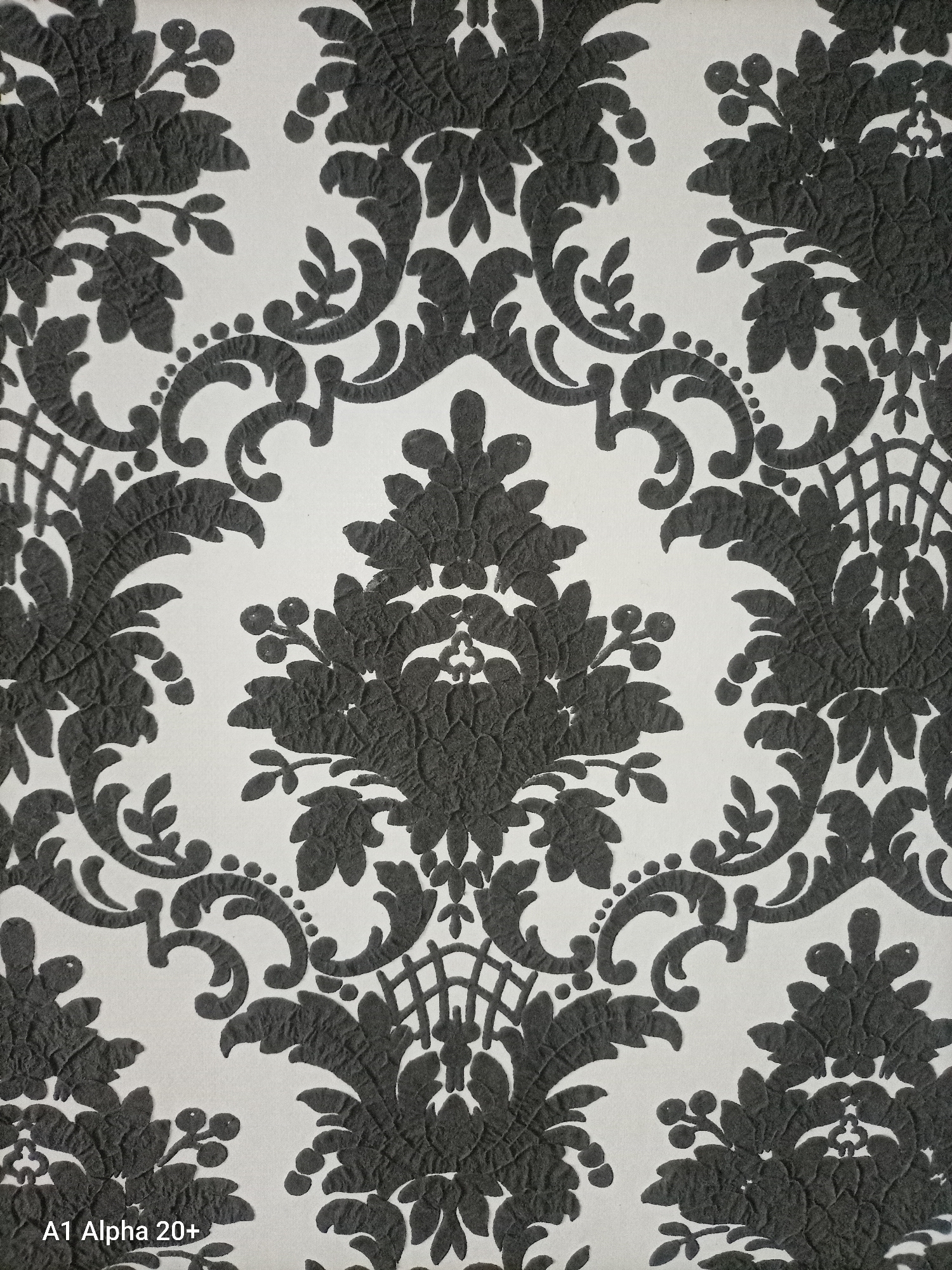 The image displays a close-up, full-frame view of a textured wallpaper with a black damask pattern against a white background. The pattern features ornate, scrolling floral motifs and foliage, with a slightly raised, embossed texture visible on the black elements. There is no discernible location in the city of Улукаўскі сельскі Савет, Belarus, as the image focuses solely on the wallpaper pattern. The composition is static and does not depict any people, actions, or interactions. The lighting is even, highlighting the texture of the wallpaper. A small, partially visible text overlay at the bottom left corner reads "A1 Alpha 20+".