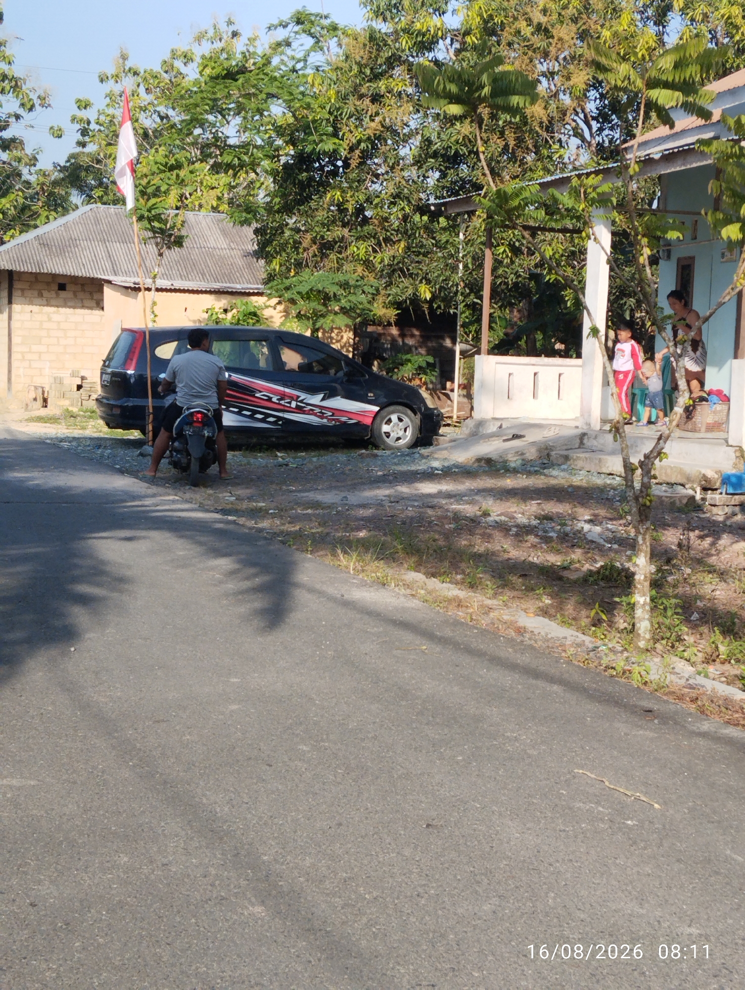 In an outdoor scene, a man is seated on a motorcycle on the side of a paved road. He is wearing a gray t-shirt and dark shorts. The motorcycle is dark-colored with some red and white decals on the side. Behind the motorcycle is a dark-colored SUV with a red, white, and black stripe decal. A red and white flag is flying from a pole next to the SUV. To the right of the SUV, there is a light blue house with a covered porch. Two children and an adult are standing on the porch. One child is wearing a red and white outfit, and the other is in a blue shirt and shorts. The adult, presumably a woman, is wearing a patterned dress. A laundry basket and other items are visible on the porch. The environment is characterized by trees and vegetation. The road surface is asphalt, with some gravel and dry grass on the shoulder. Shadows of trees are cast on the road. The date and time stamp "16/08/2026 08:11" is visible in the lower right corner of the image.